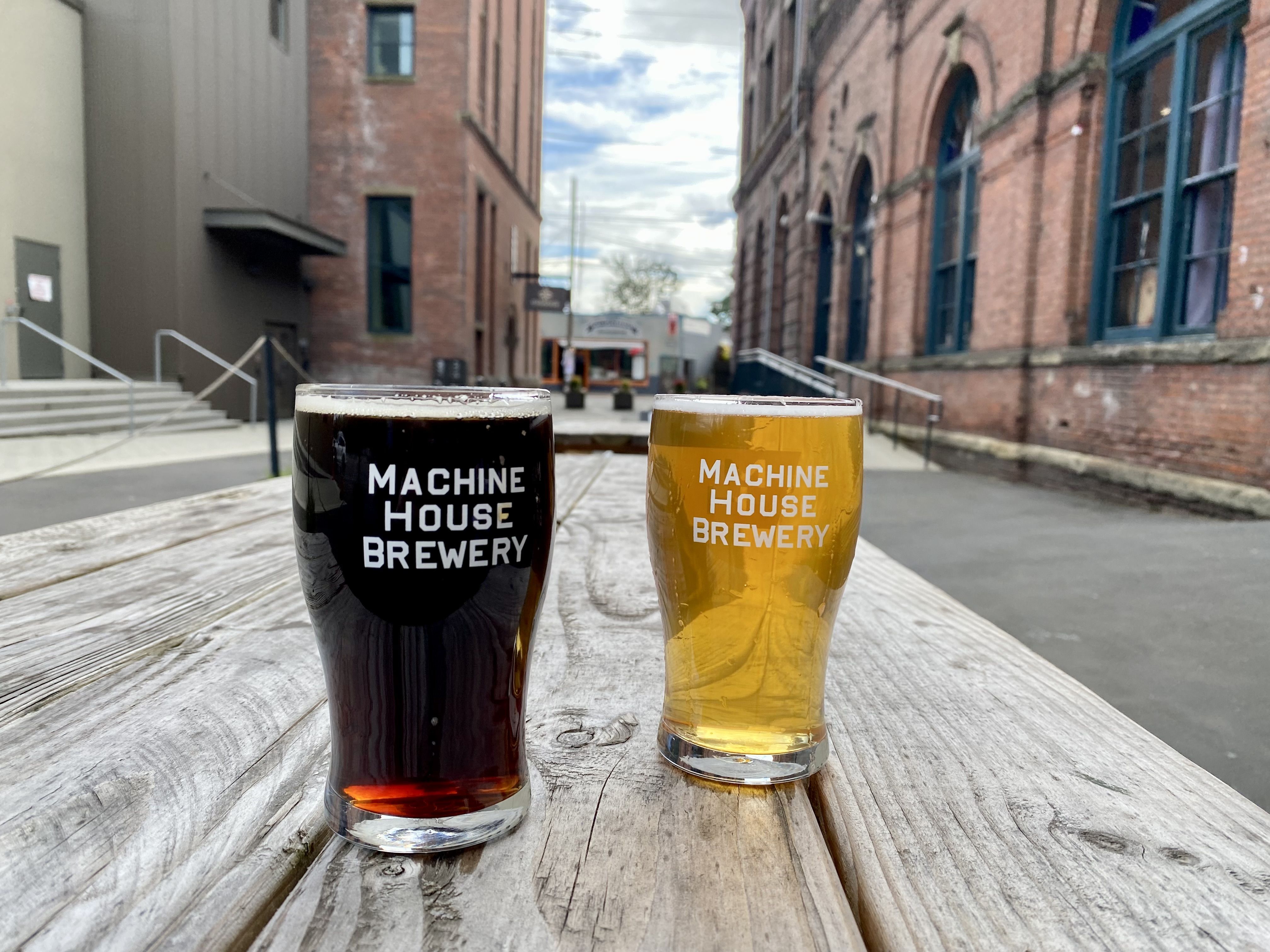 Two beers, one dark and one light, in glasses that say Machine House Brewery on a wooden picnic table with an open space between two brick buildings in the background. 