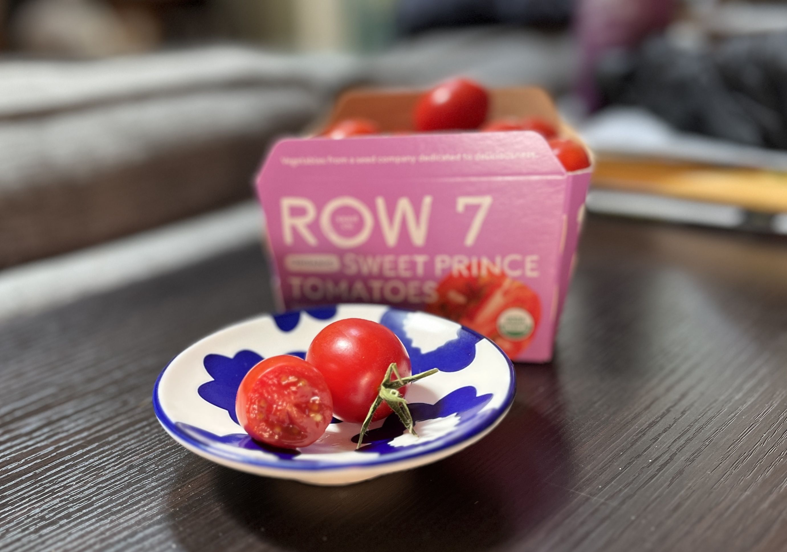 Two red cherry tomatoes, one whole and one cut in half, on a white plate with blue floral patterns; a pink container labeled "ROW 7 SWEET PRINCE TOMATOES" is in the background on a dark wood table.