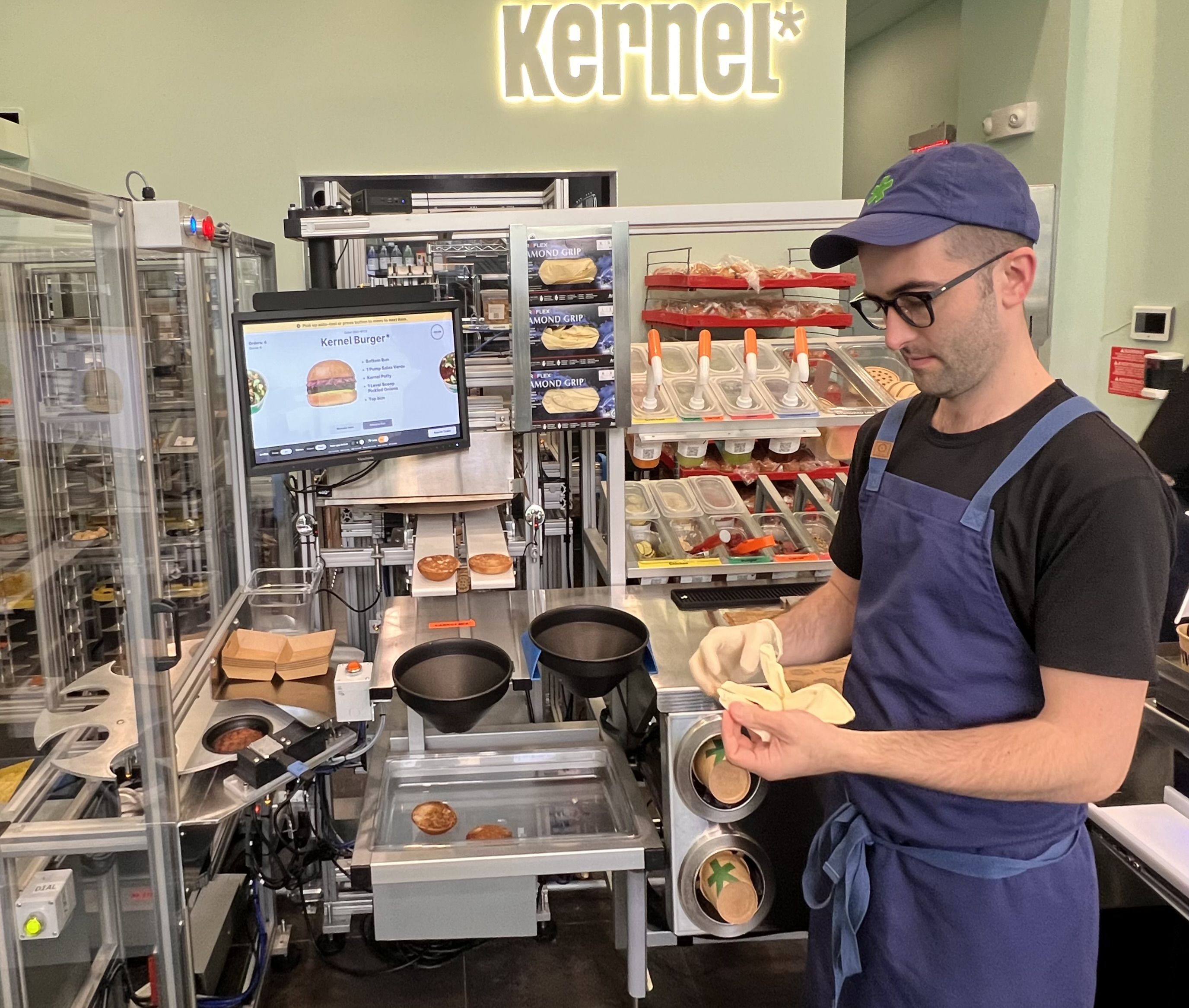 The chef at Kernel stands next to rows of ingredients and a screen displaying a burger.