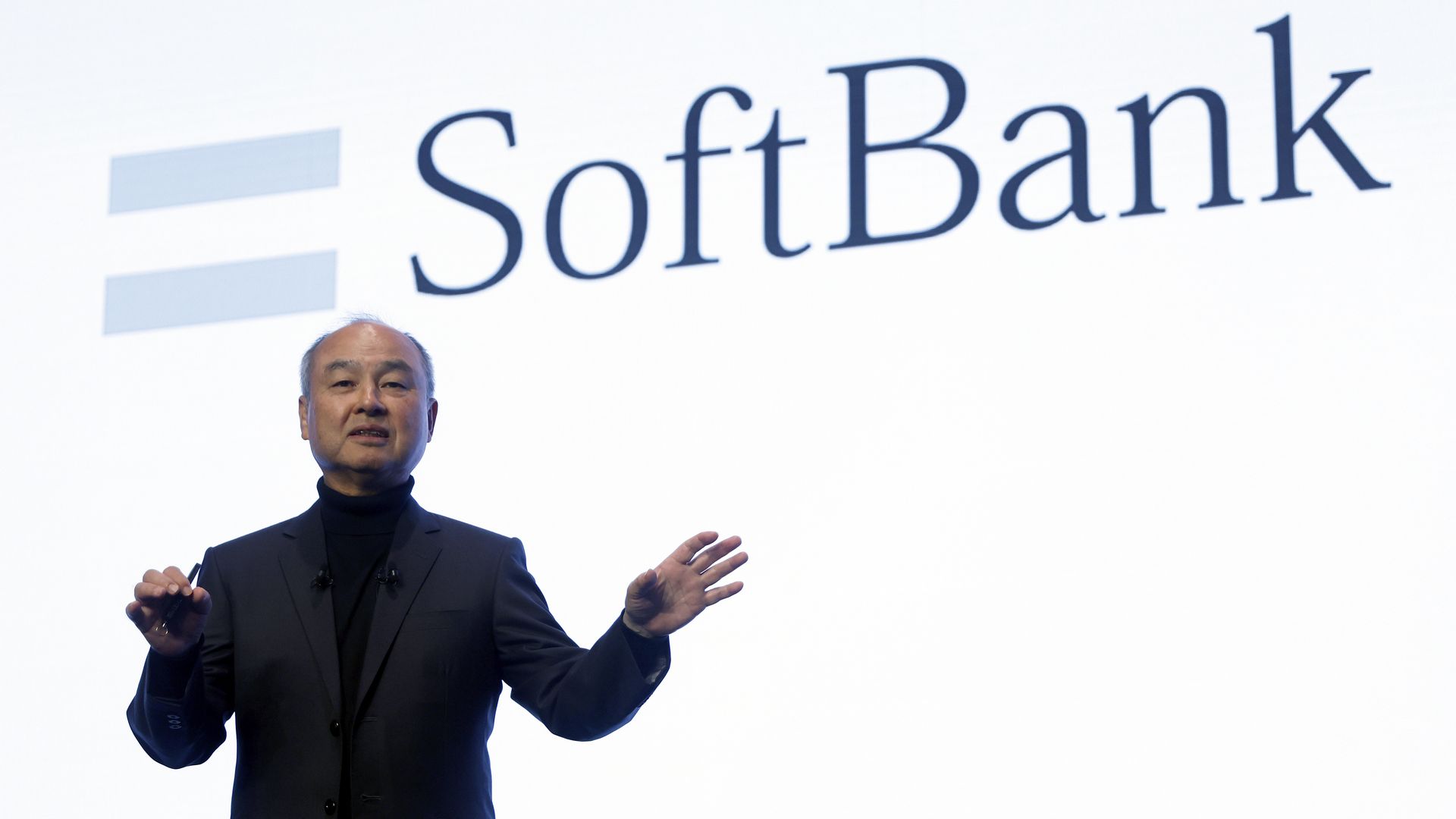 Person in black suit and turtleneck speaking on stage with a large SoftBank logo projected behind them on a white background.