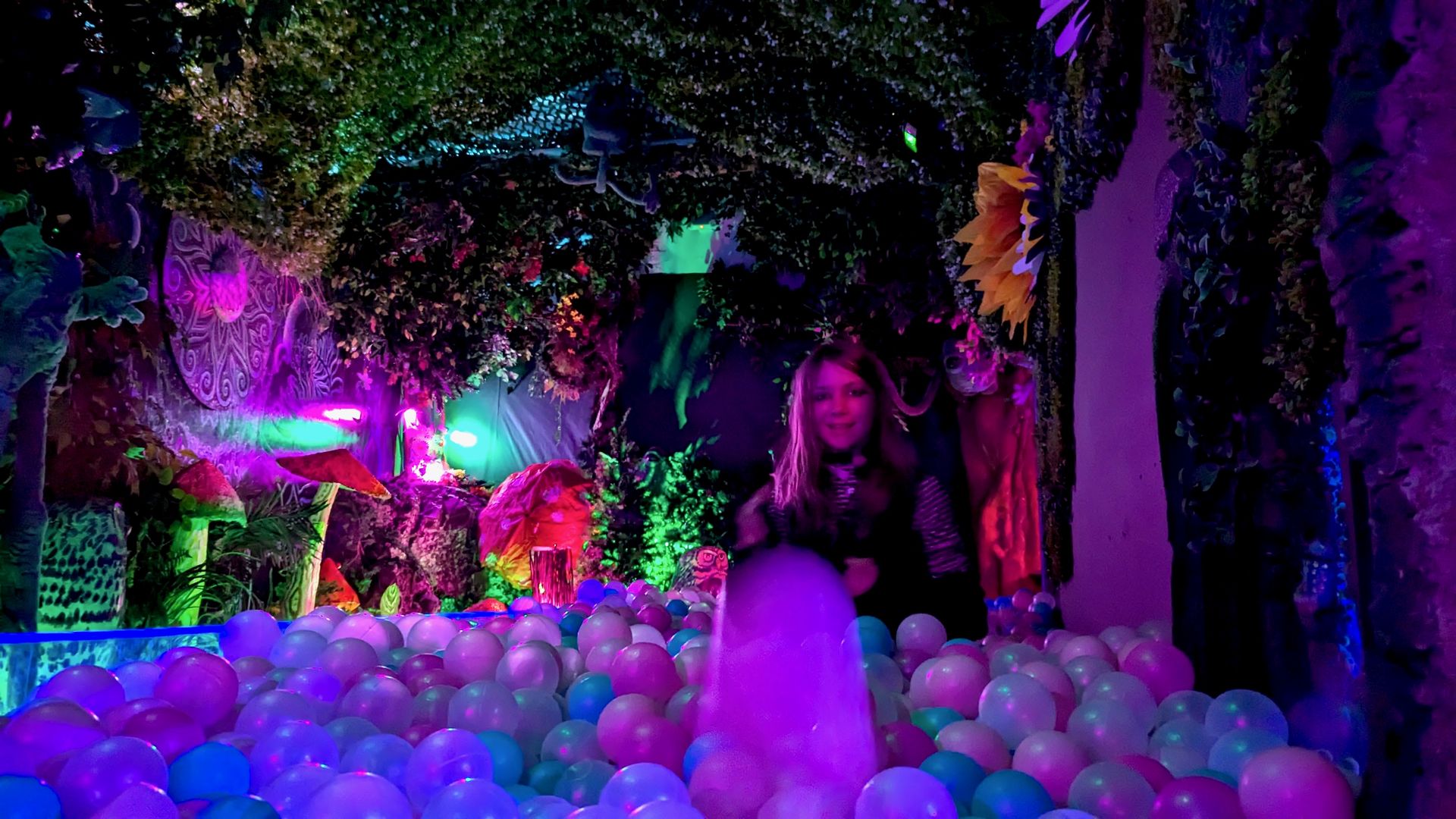 A child throws a ball in a ball pit with a colorful fantasy swamp scene in the background, with lighted mushrooms and plants.
