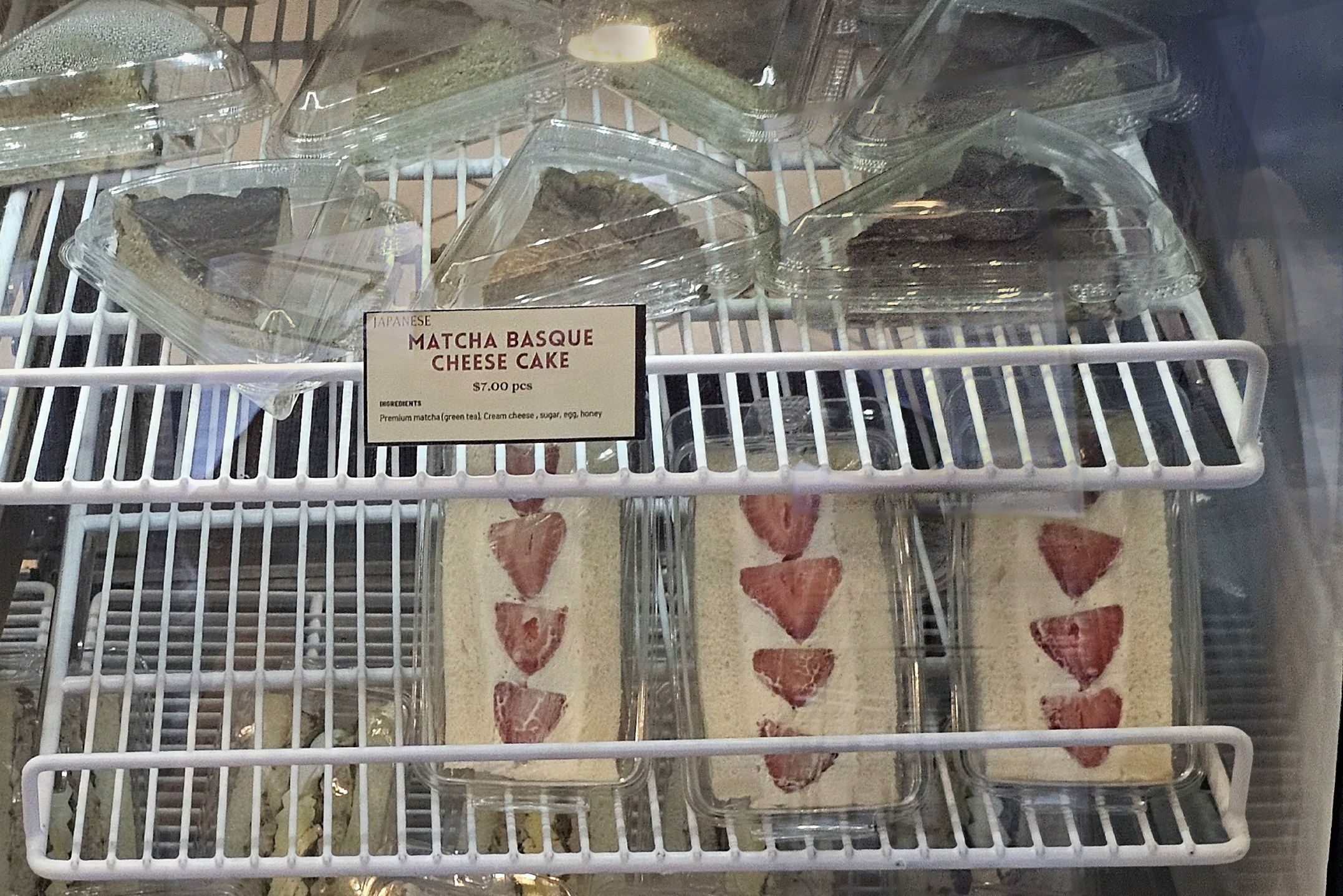 Bakery display case with plastic-wrapped trays of matcha Basque cheese cake slices; a sign reads "Japanese Matcha Basque Cheese Cake" $7.00 per piece.