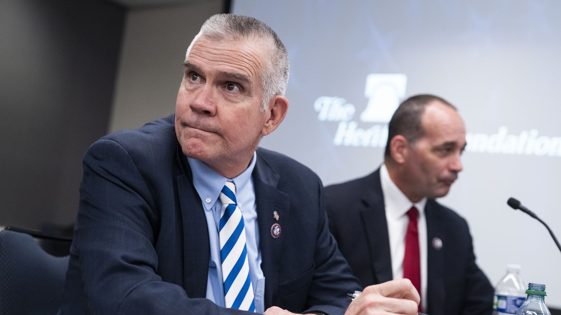 Rep. Matt Rosendale, wearing a dark blue suit jacket, light blue shirt and blue-and-white striped tie, seated at a table with bottles of water next to Rep. Bob Good.