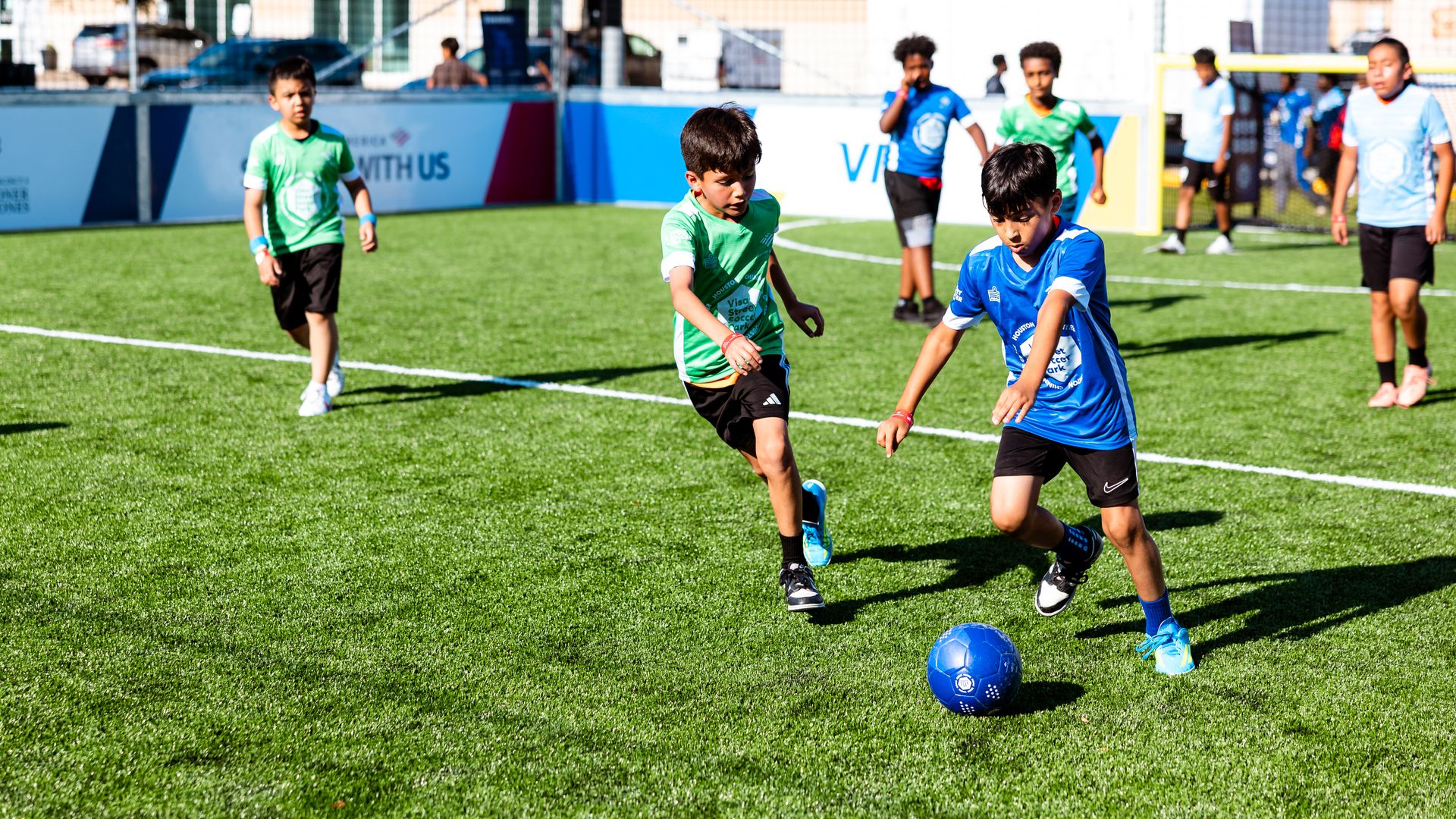Youth soccer game on a bright green artificial turf field. Boys in green and blue jerseys chase a blue ball; a small crowd and banners line a fence in the background near a goal.