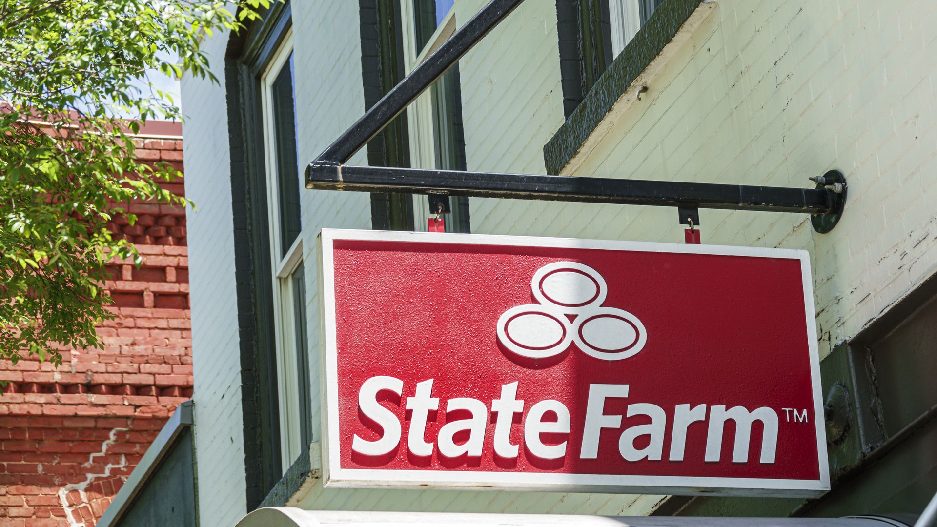 Red and white State Farm sign with logo of three interconnected ovals, hanging outside a light green building with black-trimmed windows and nearby green tree branches.