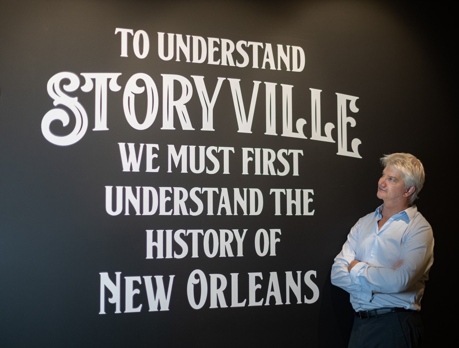 A man crosses his arms and stands next to a panel that reads "To understand the history of Storyville, we must first understand the history of New Orleans."