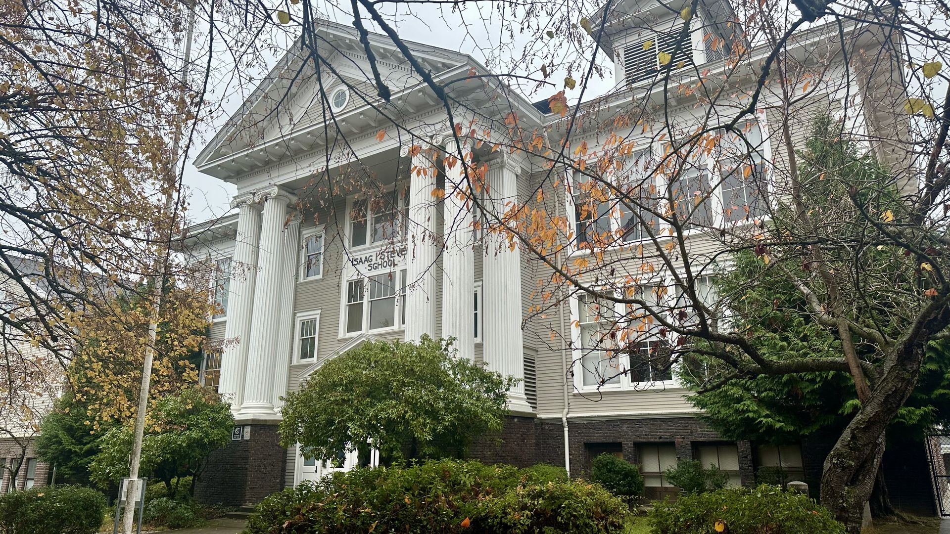 A view of a school with a classical-style portico and pediment, with bushes in the foreground and bare tree branches reaching in from outside the photo frame.