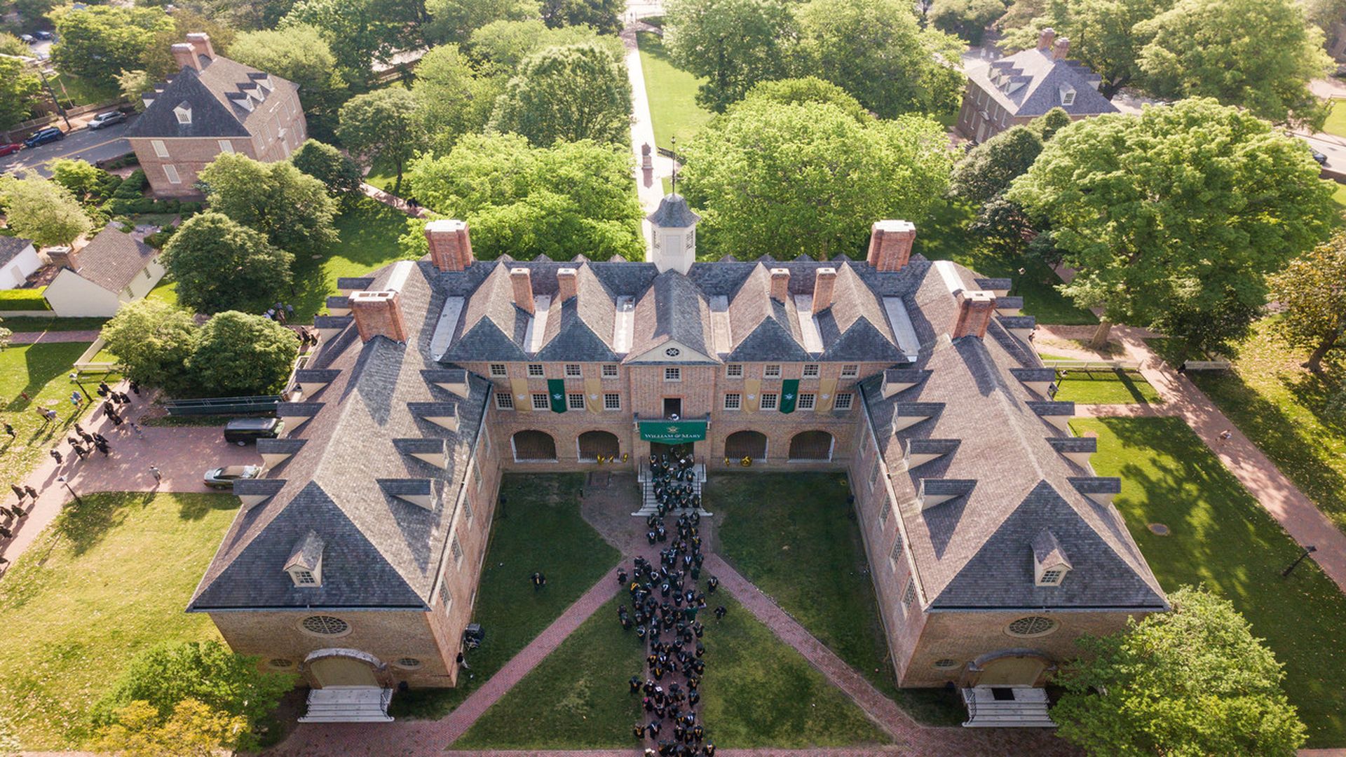 Aerial view of a U-shaped brick university building with slate roofs, lush trees, and a line of graduates walking toward the central entrance on a manicured lawn.
