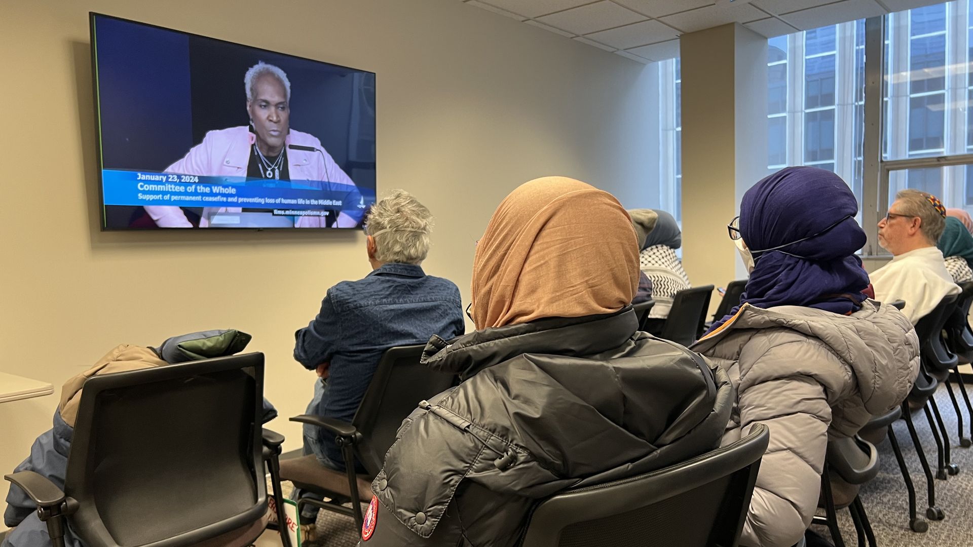 Two women in head-coverings face away from the camera and watch a television screen displaying a woman in a pink blazer.