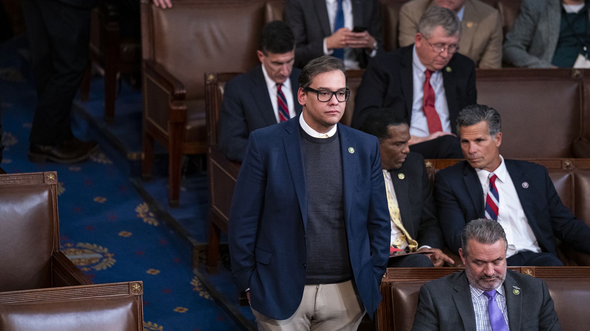 Rep. George Santos, wearing glasses, a gray suit over a white shirt and a blue suit jacket, stands on the Republican side of the House chamber.