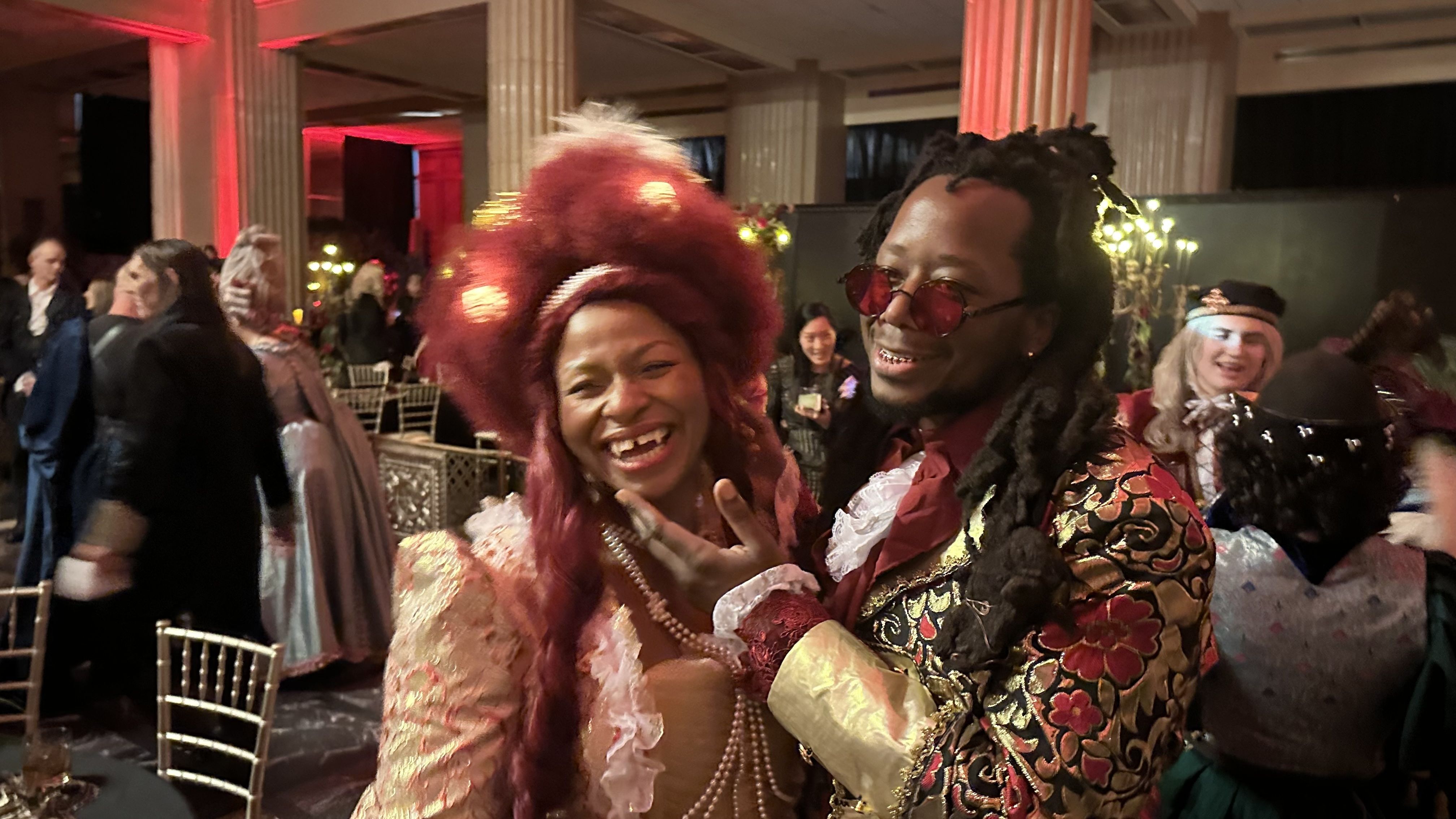 Two people at a lively party in ornate, colorful 18th-century style costumes; woman with large red hair and pearls, man in floral gold jacket and red-tinted glasses, smiling warmly.