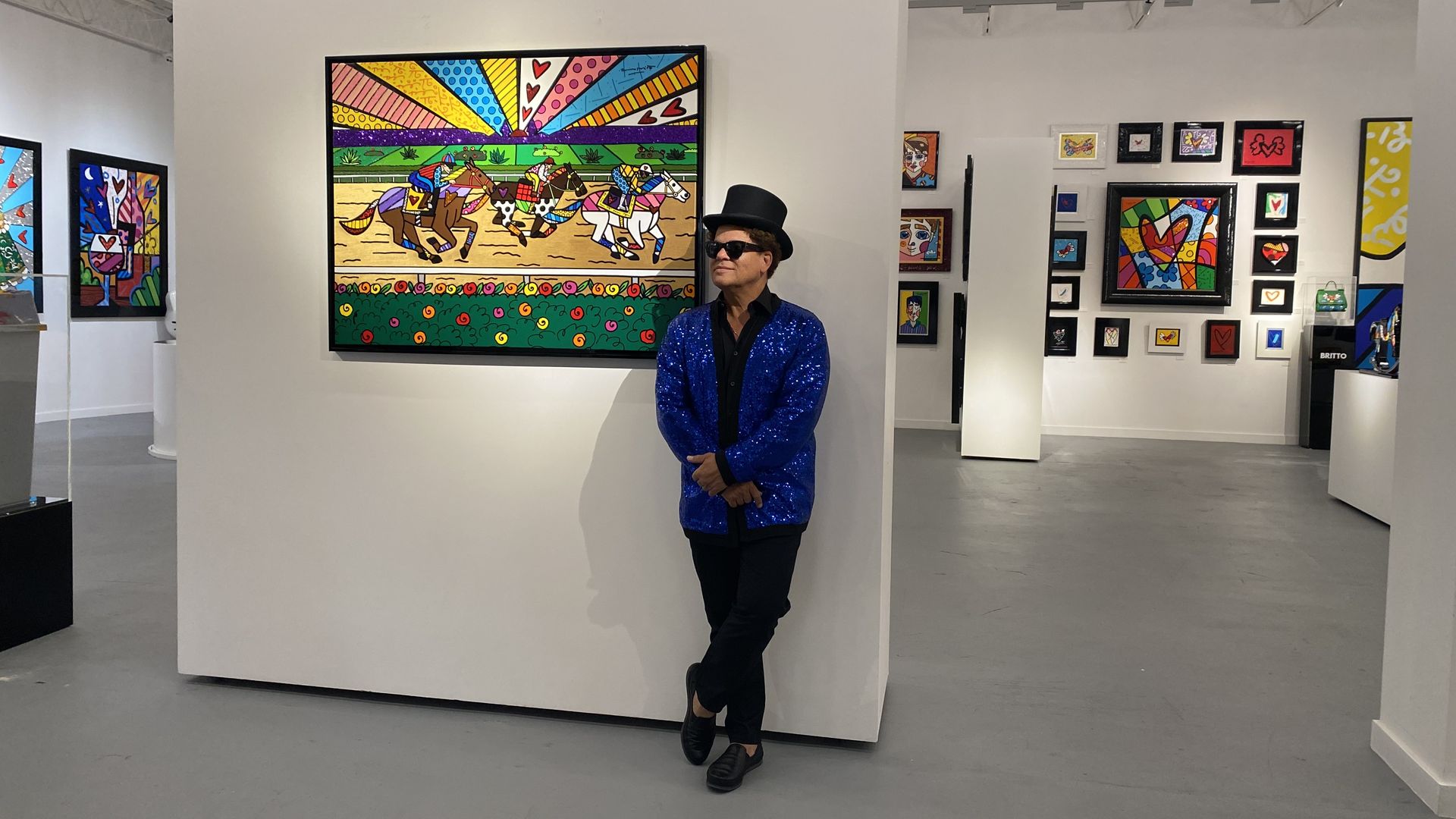 Romero Britto wears a blcak top hat and glittery blue jacket as he stands next to a painting. 