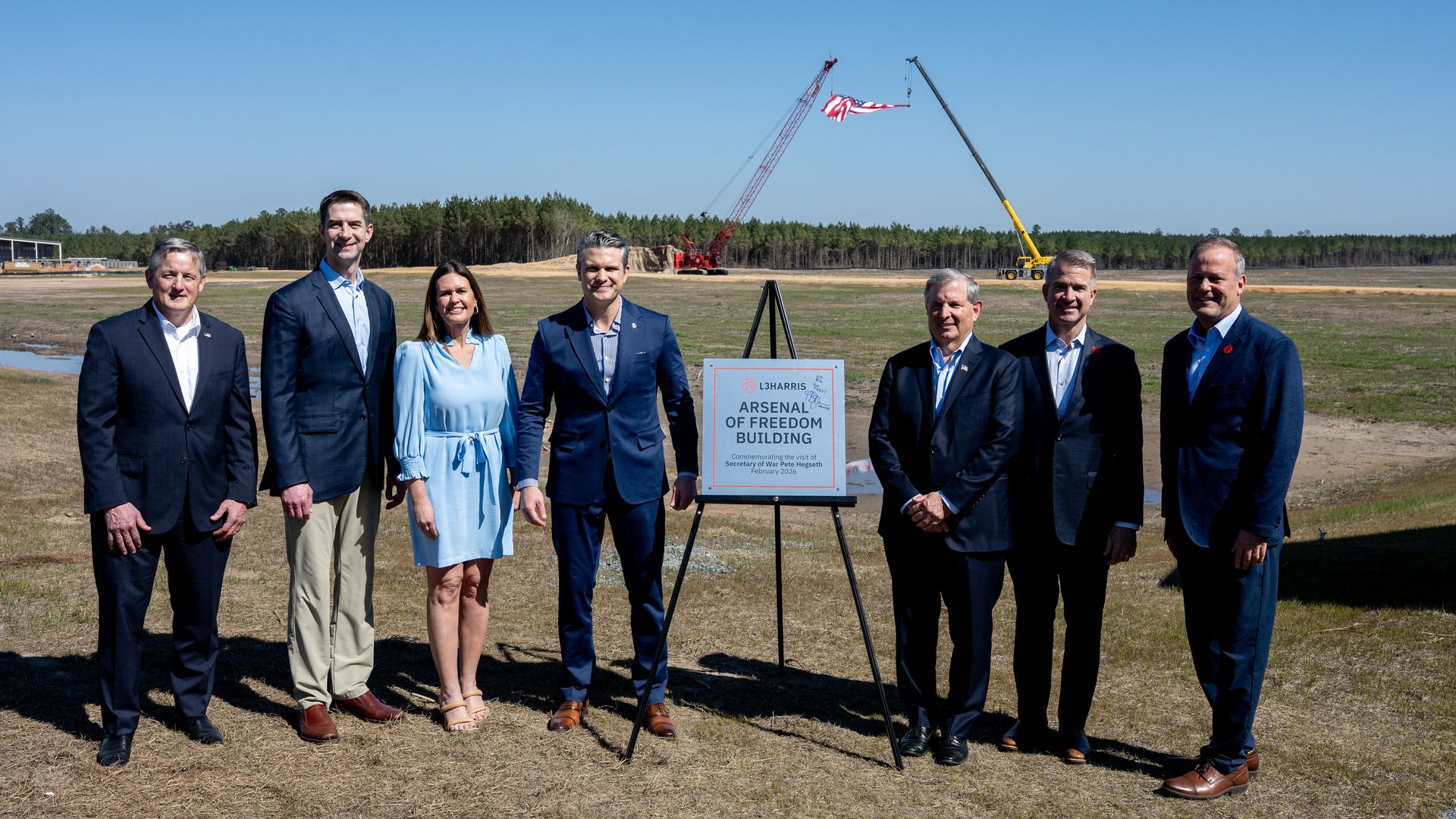 Seven people in business attire stand outdoors on grass near a sign reading "Arsenal of Freedom Building," with two cranes holding an American flag in the background under clear blue sky.