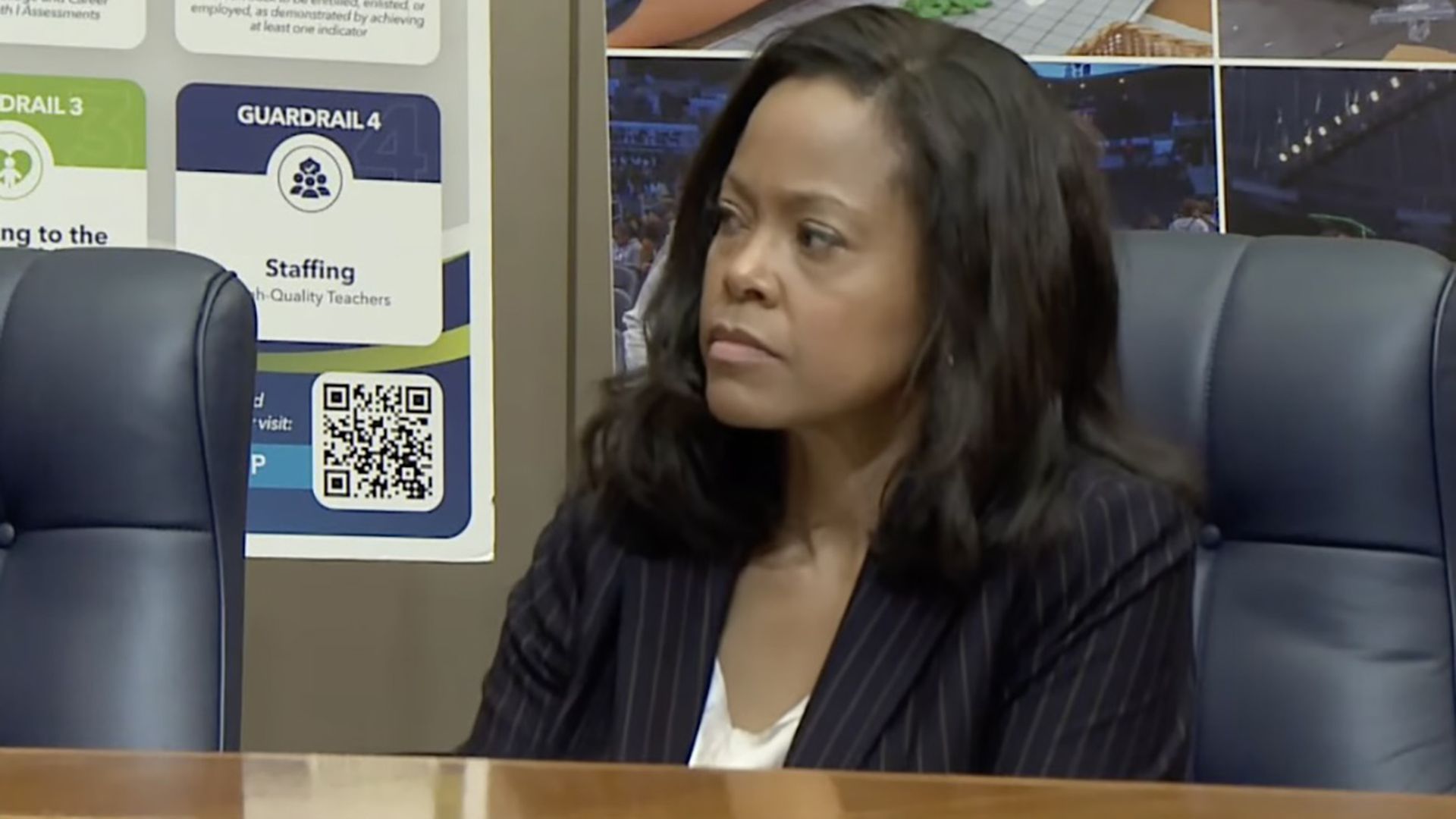 Professional woman with dark hair in a navy pinstripe blazer and white blouse sits at a conference table, looking to her left. Behind her are posters with colorful signs and a QR code.