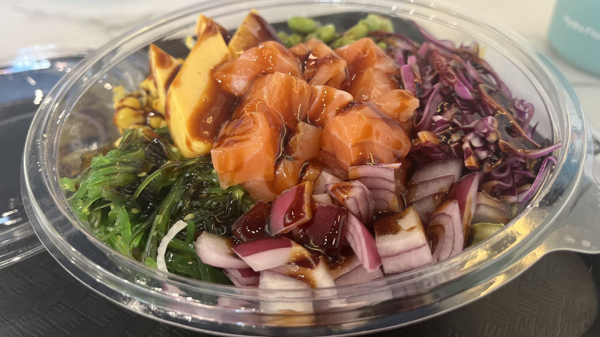 Clear plastic bowl containing a poke bowl with diced salmon, seaweed salad, red cabbage, red onions, avocado, and drizzled dark sauce on top.