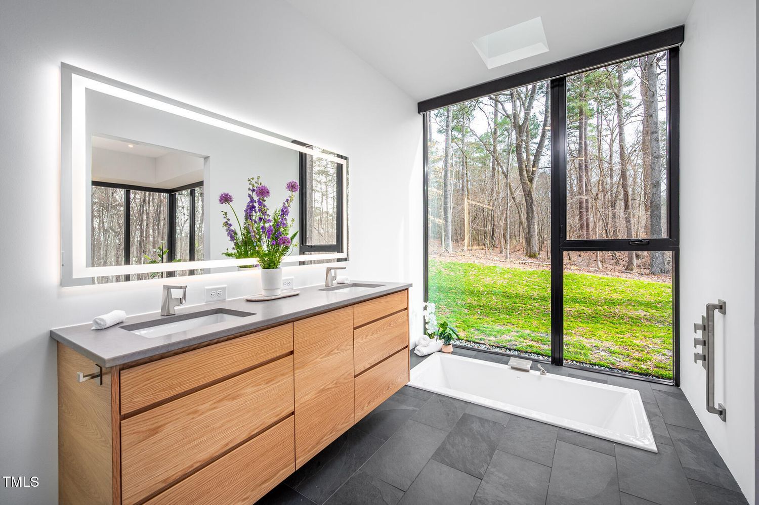 Modern bathroom with wood vanity, gray countertop, double sinks, illuminated rectangular mirror, sunken white bathtub next to large floor-to-ceiling window showing green yard and trees.