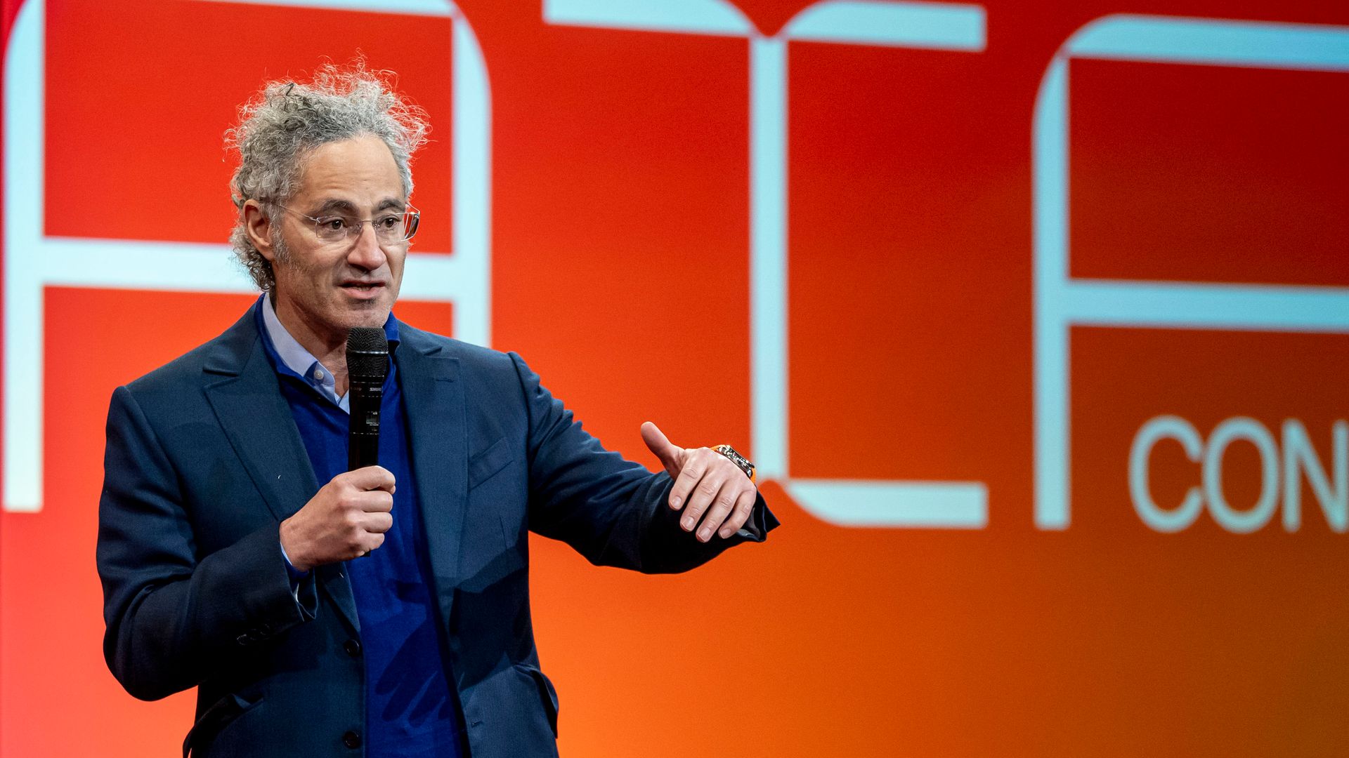 A man with a microphone in hand speaks while gesturing. 