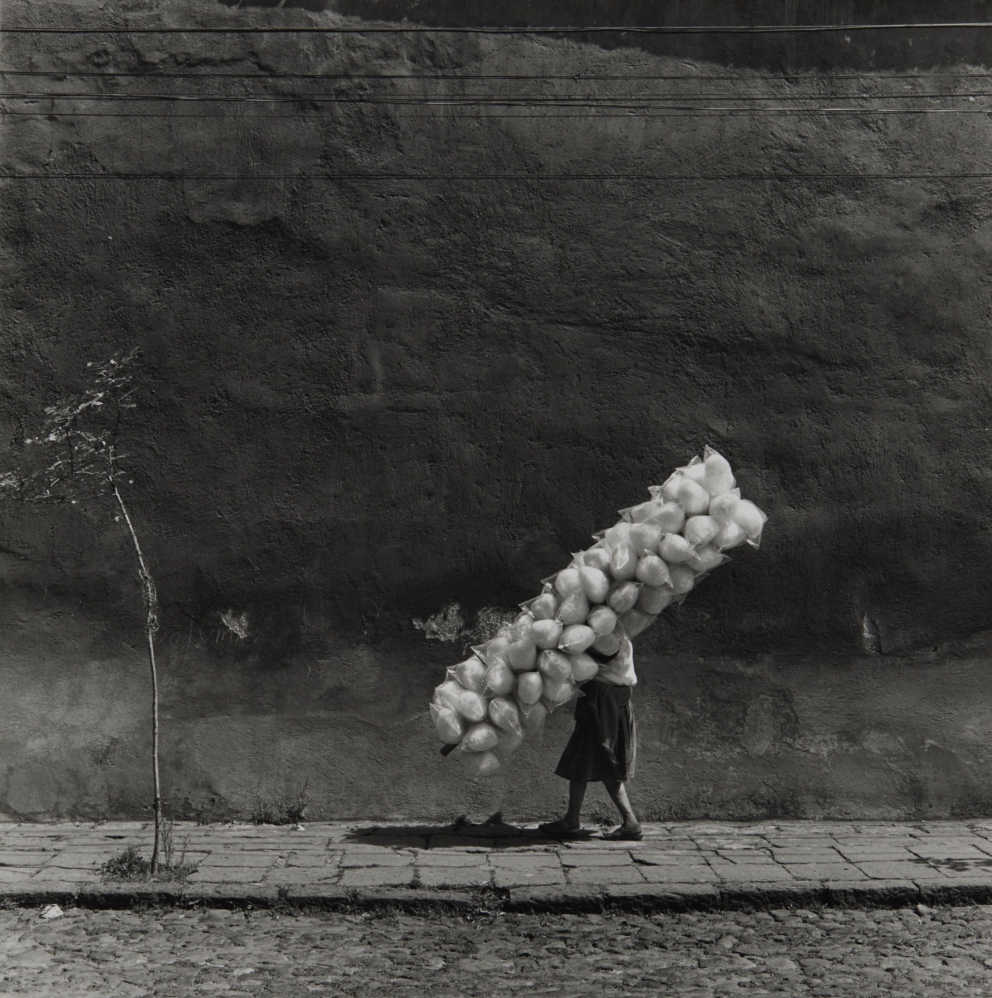 Black-and-white image of a person in a dark coat walking along a cracked sidewalk, carrying a long, stacked bundle of white round objects against a rough wall with power lines overhead.
