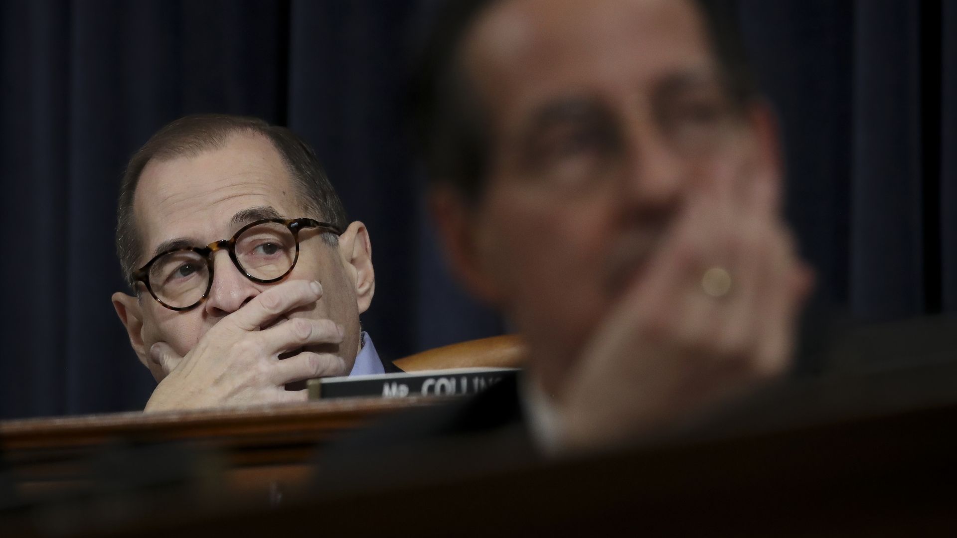 Rep. Jerrold Nadler with his hand over his mouth, sitting at a wooden dais in front of a blue curtain with a blurry Rep. Jamie Raskin in the foreground.