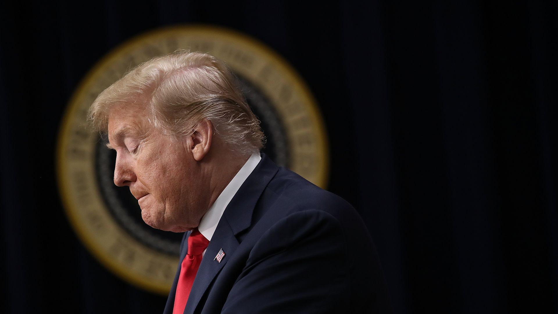 Donald Trump looks down bites his lip before a dark background.