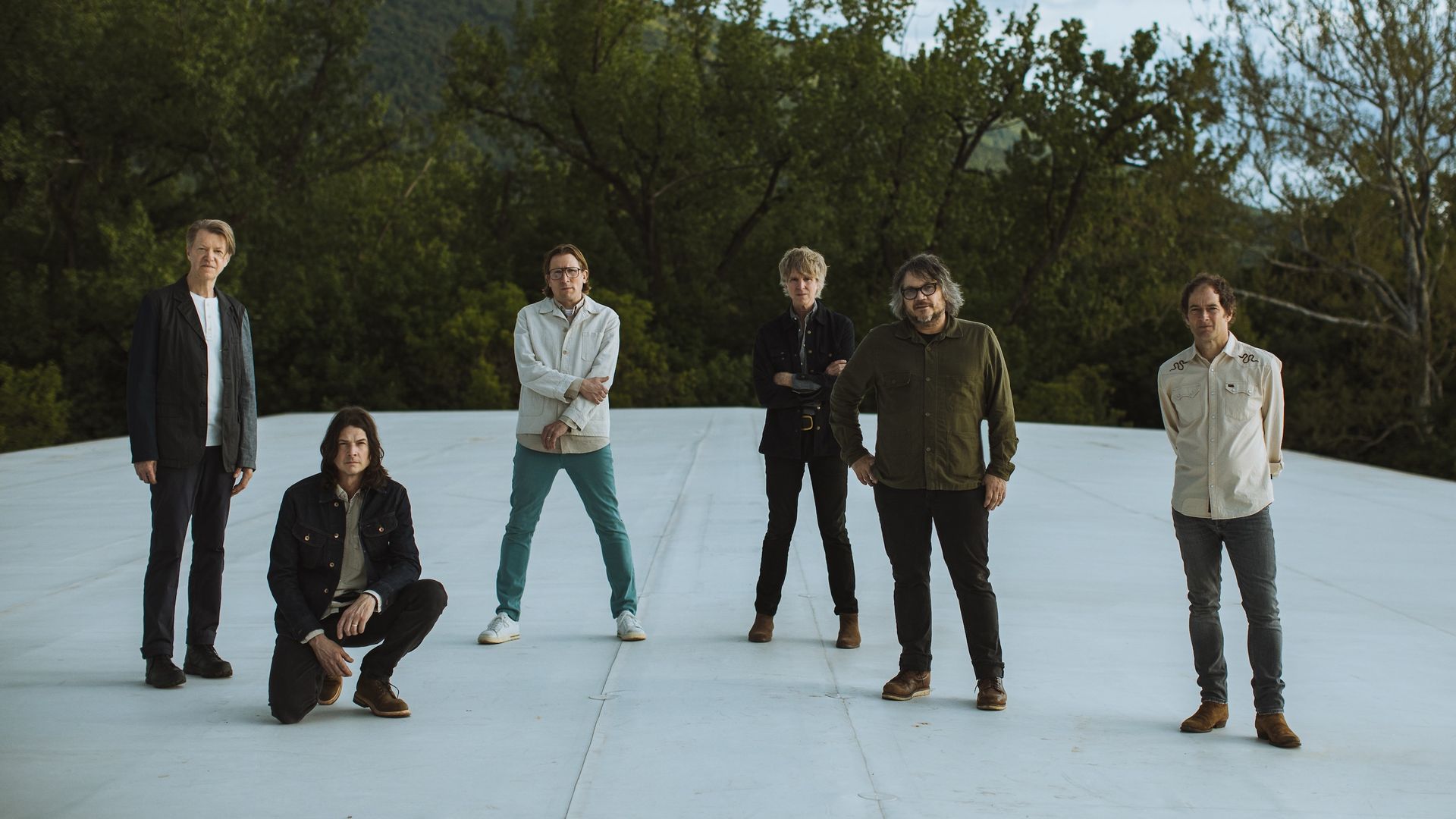 The six members of Wilco posing outdoors on a white surface with green trees and a mountain in the background, wearing casual clothing in various colors including black, white, and green.