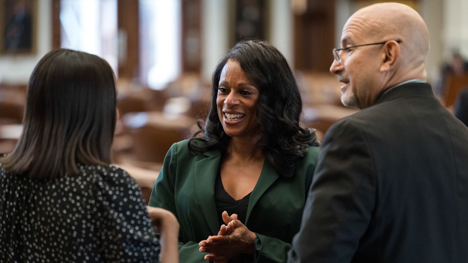 Three people in conversation in a room with brown chairs; a smiling woman in a green blazer faces a woman with dark hair in a black and white polka dot top, and a bald man in glasses in a black jacket.