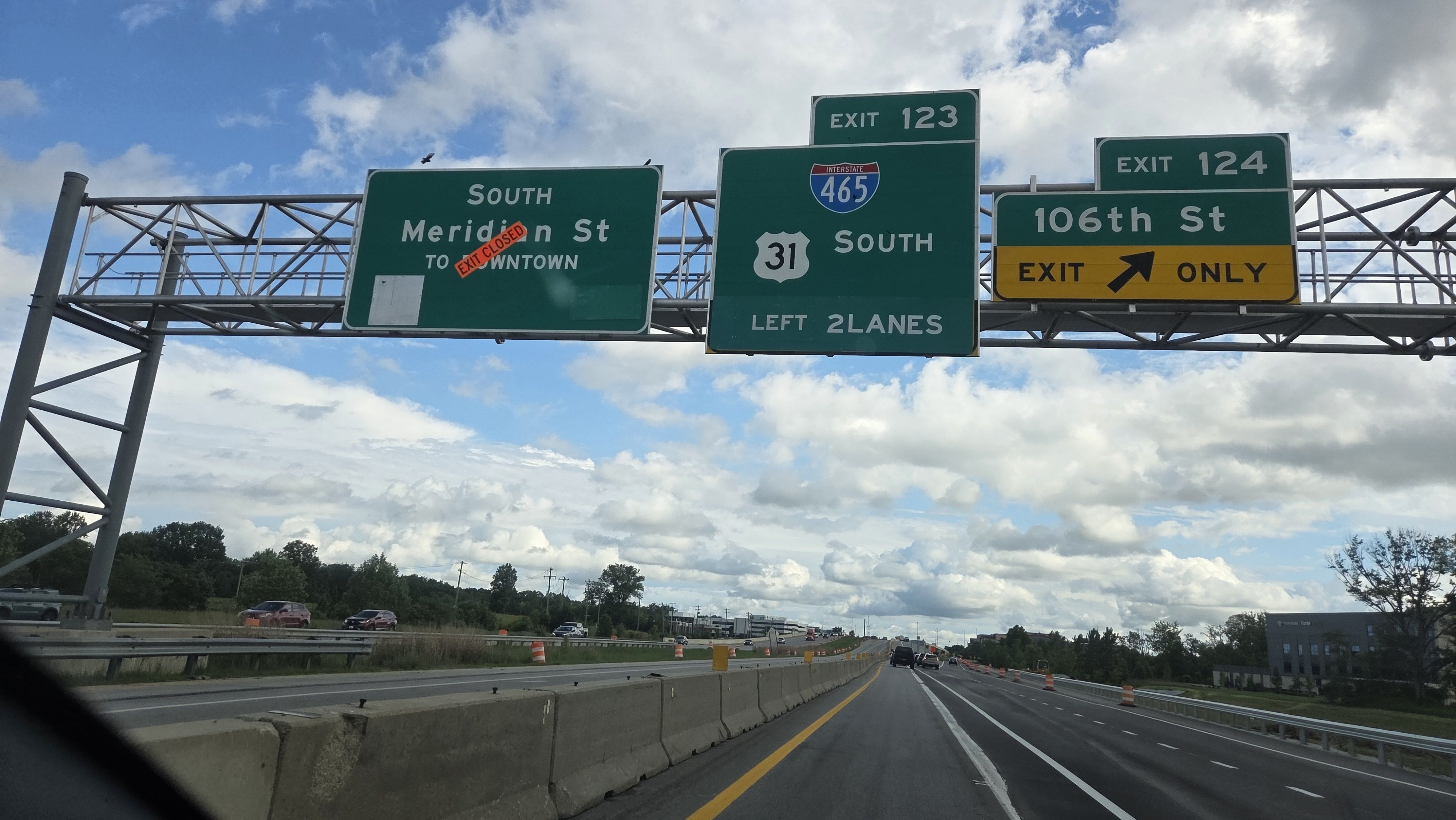 Three green highway signs overhead on a sunny day: Meridian St South - EXIT CLOSED; I-465 South with shield 31; Exit 124 for 106th St with yellow EXIT ONLY panel; concrete barrier and traffic.