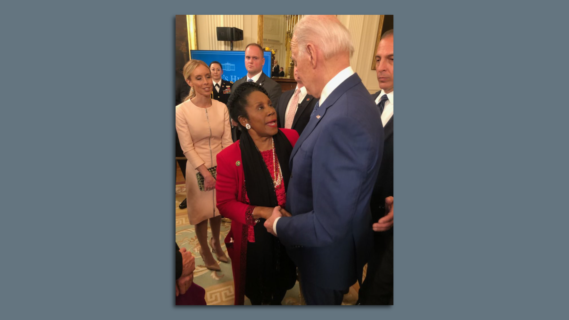 Rep. Sheila Jackson Lee, wearing a red jacket and black scarf, looks up toward President Joe Biden as the two speak with one another. 