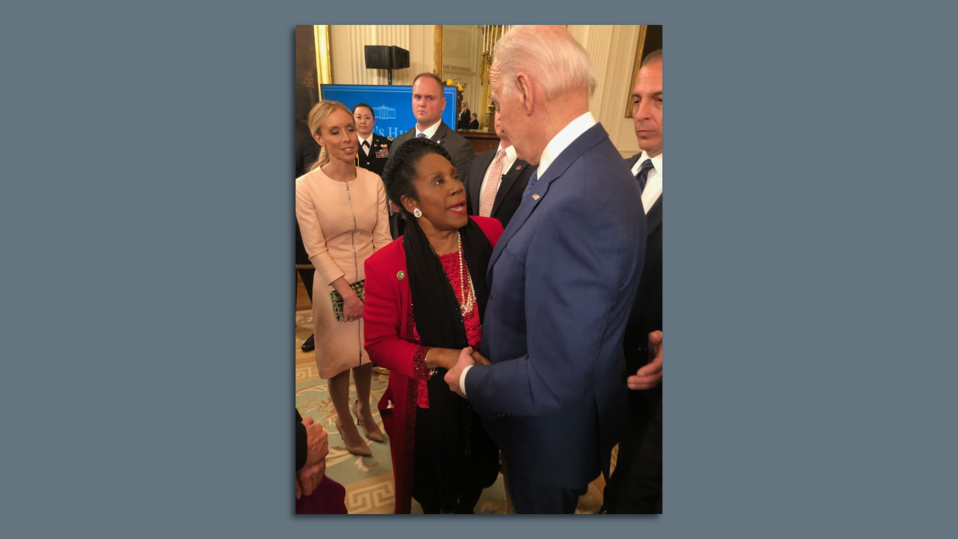 Rep. Sheila Jackson Lee, wearing a red jacket and black scarf, looks up toward President Joe Biden as the two speak with one another.