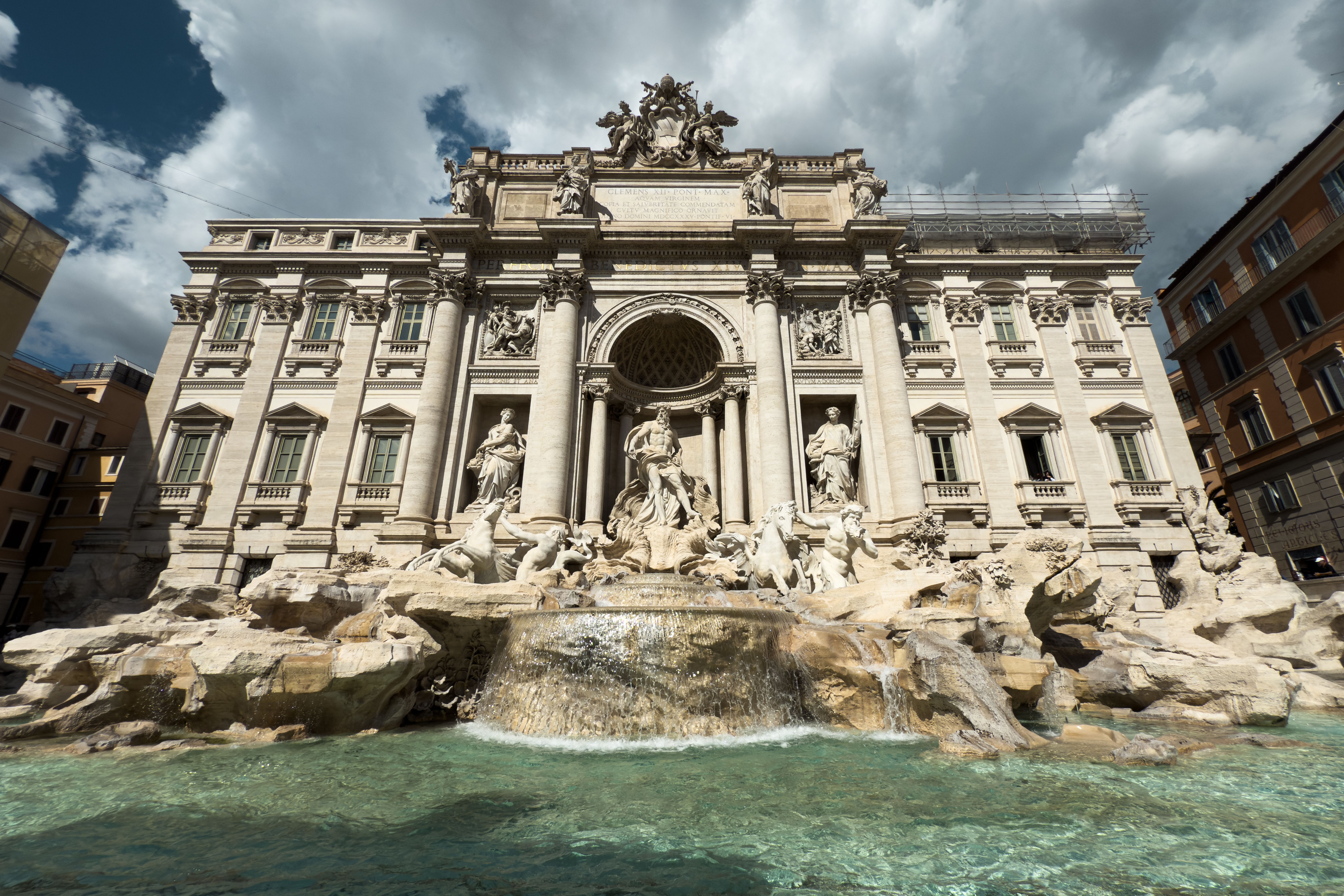 The Trevi Fountain, a large ornate fountain with statues of mythological figures and horses in front of a grand building under a partly cloudy sky, with clear turquoise water in the basin.