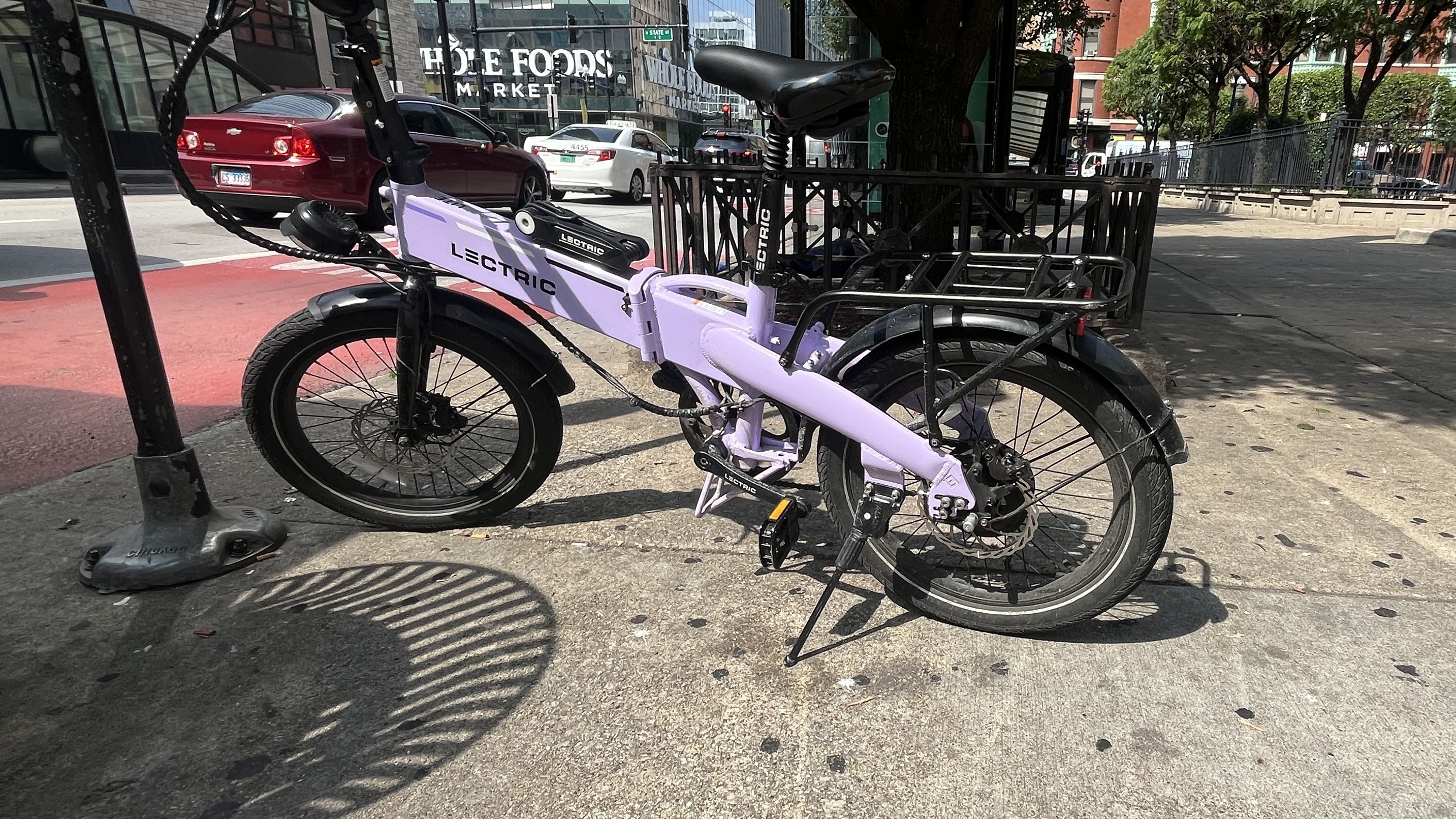 A lavender Lectric electric folding bike parked on a city sidewalk near a Whole Foods Market, with a red car and trees in the background.