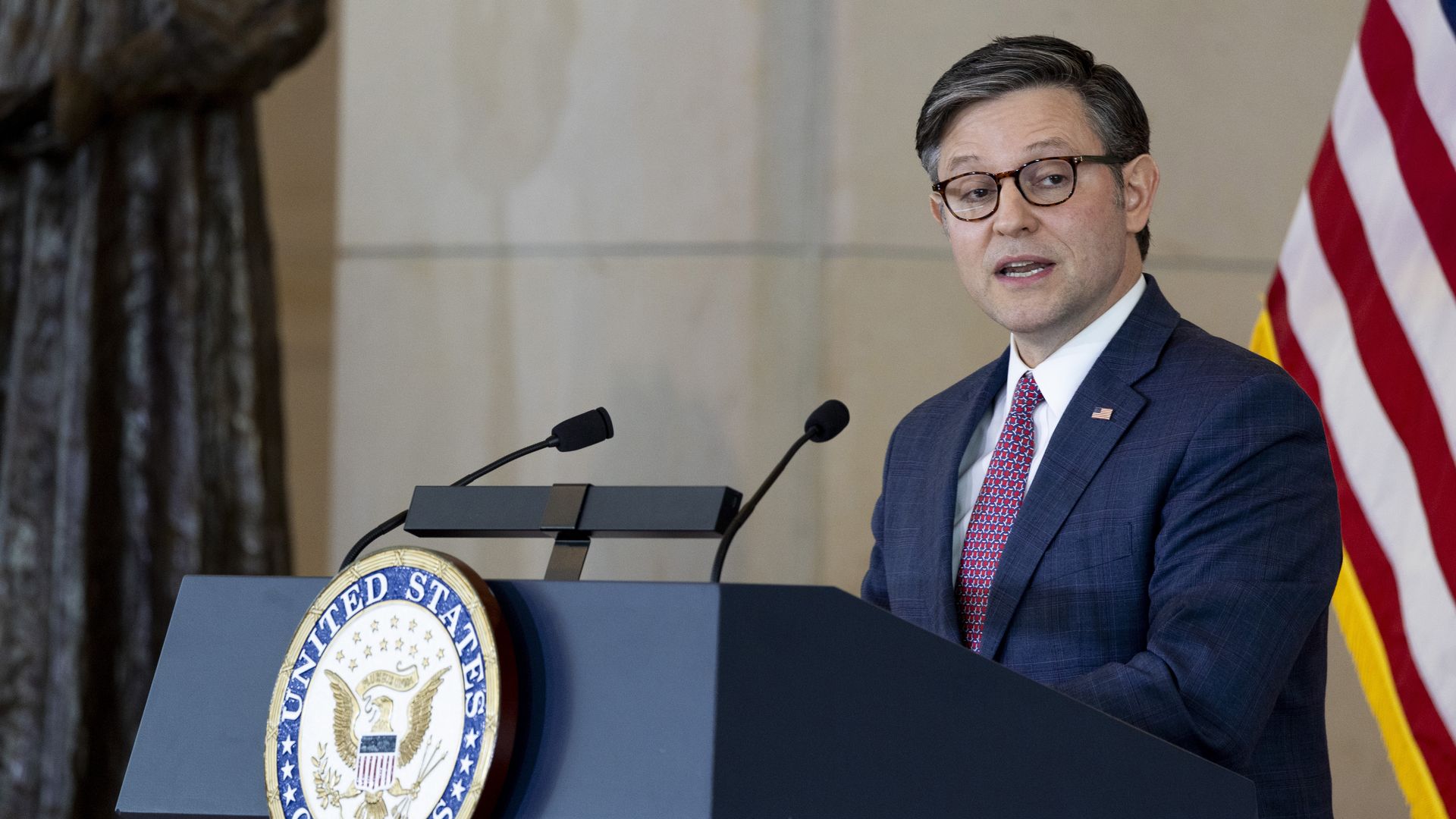 House Speaker Mike Johnson, wearing a blue suit, white shirt, red tie and glasses, standing at a podium in front of an American flag and a sandstone brick wall.