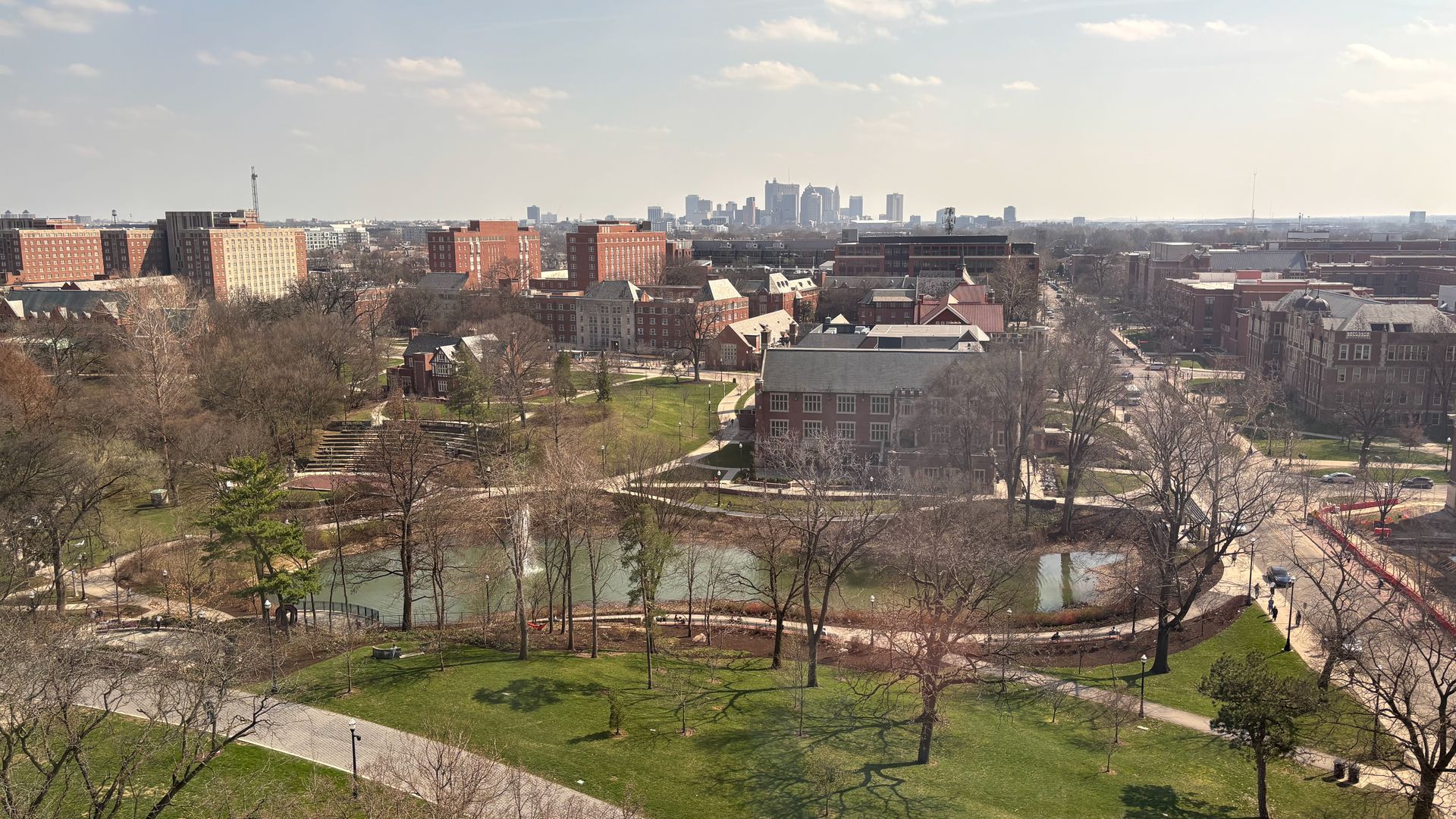 Panoramic view of Ohio State University's south campus on a sunny day: green lawns, bare trees, winding paths, and a pond with a fountain. Red-brick buildings line the mid-ground; distant skyline with tall towers.