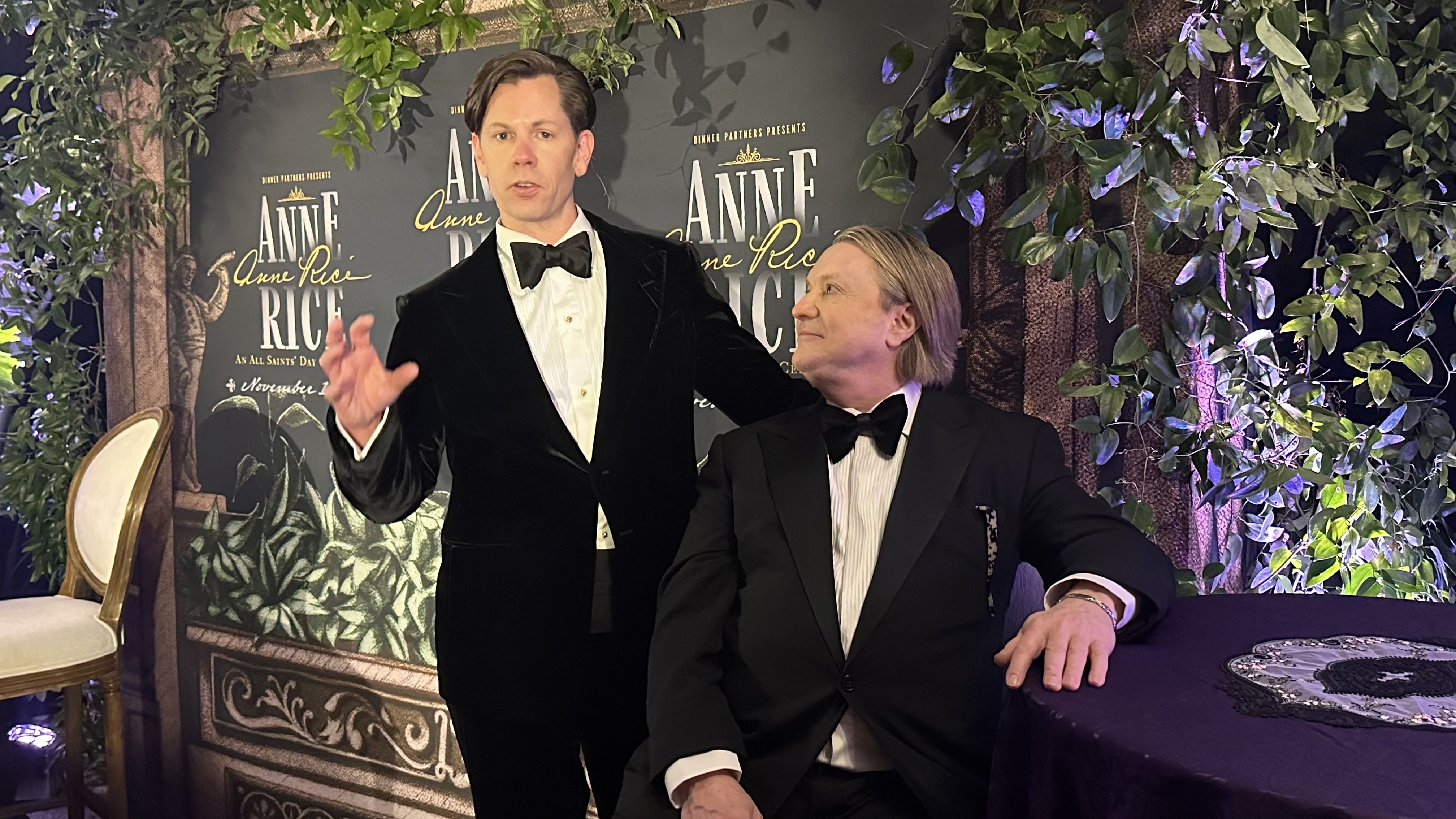 Two men in black tuxedos at an event for "Anne Rice." One stands speaking with a hand gesture, the other sits looking up at him. Green foliage and event posters in the background.