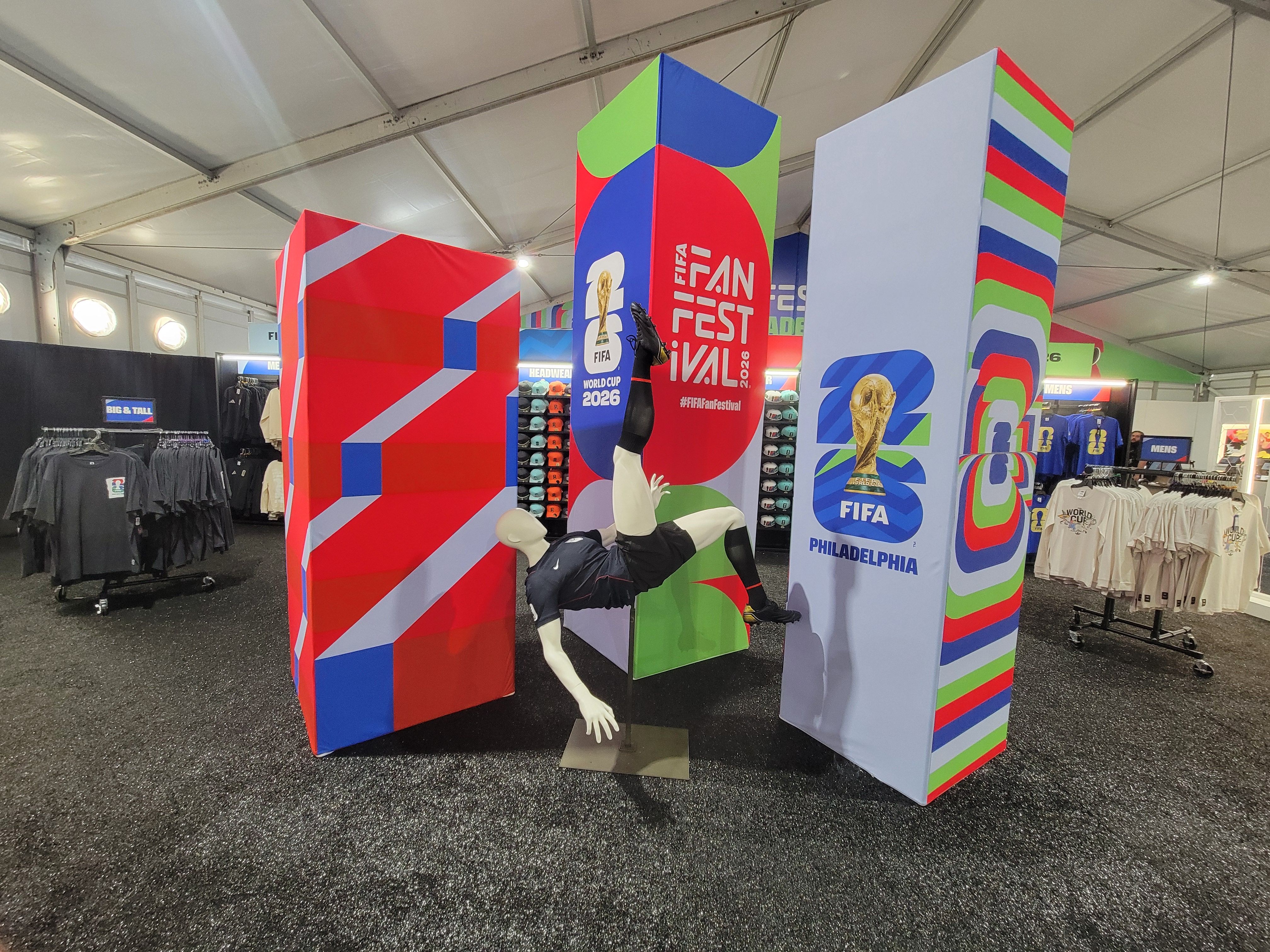 Inside a large tent, tall colorful pillars display "FIFA FAN FESTIVAL 2026" and "PHILADELPHIA," with a mannequin arm/leg at the center and clothing racks visible in the background.