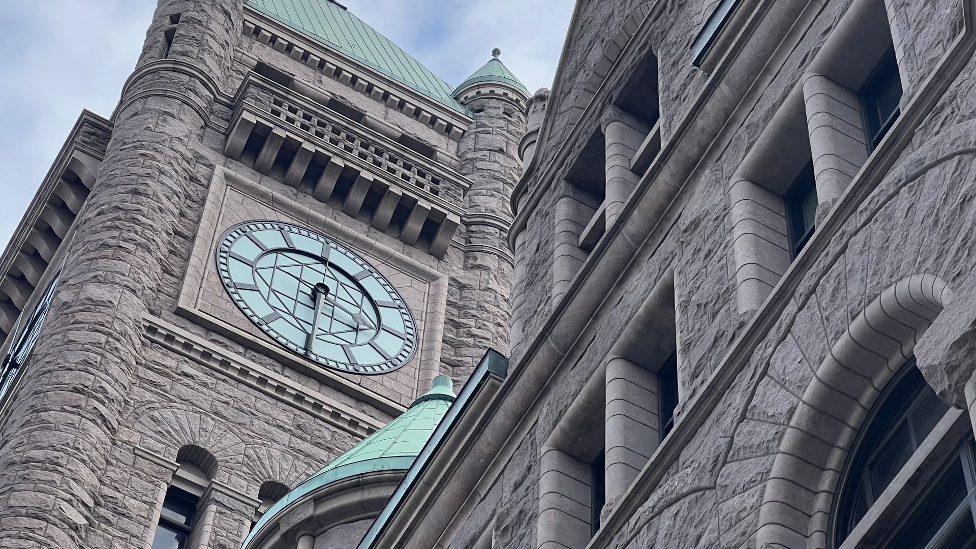 Upward view of a stone clock tower at Minneapolis City Hall with teal-green peaked roofs and turrets under a partly cloudy sky.