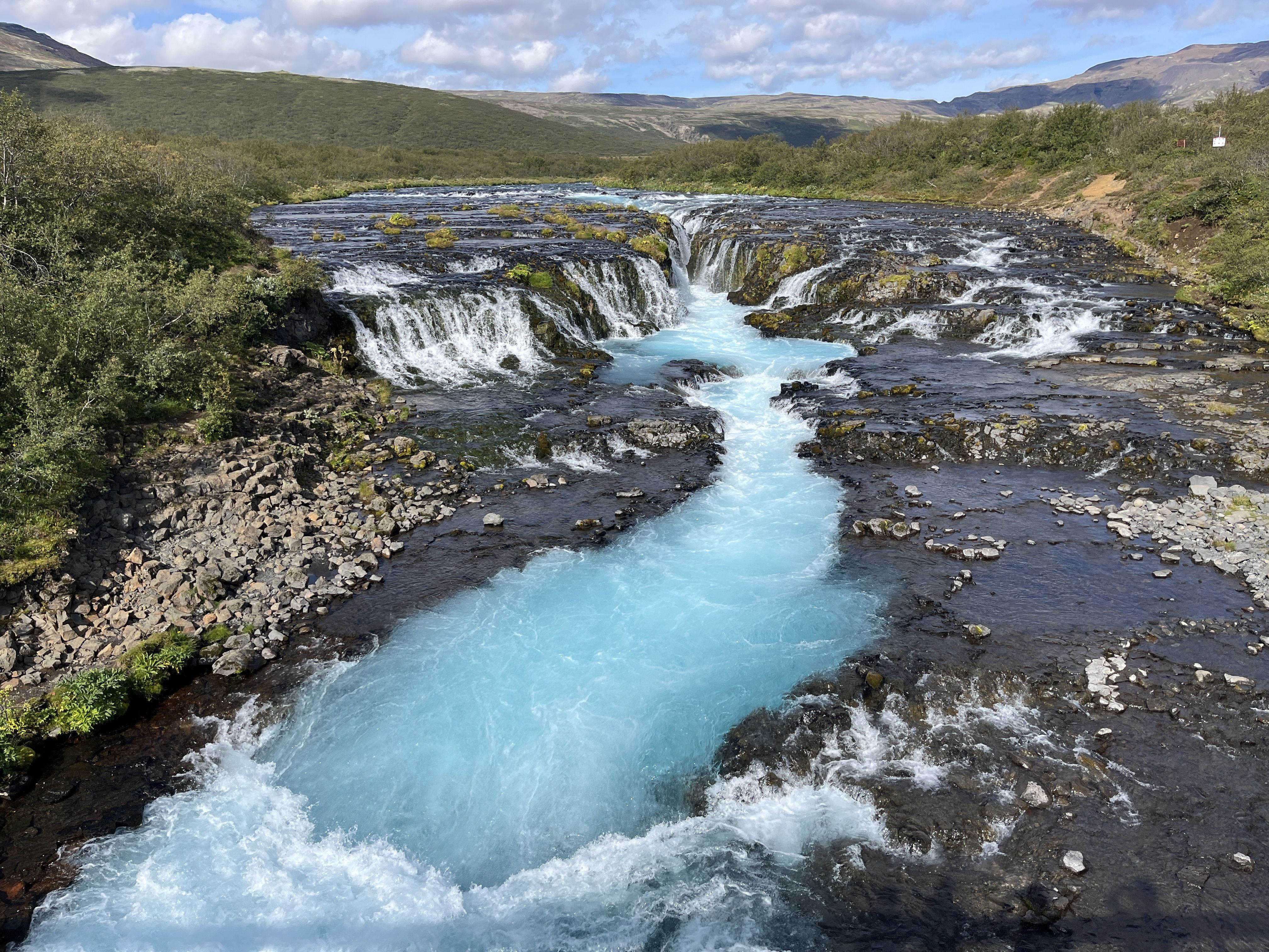 A bright turquoise river flows through dark rocky terrain with small waterfalls surrounded by green bushes under a partly cloudy blue sky and distant hills.