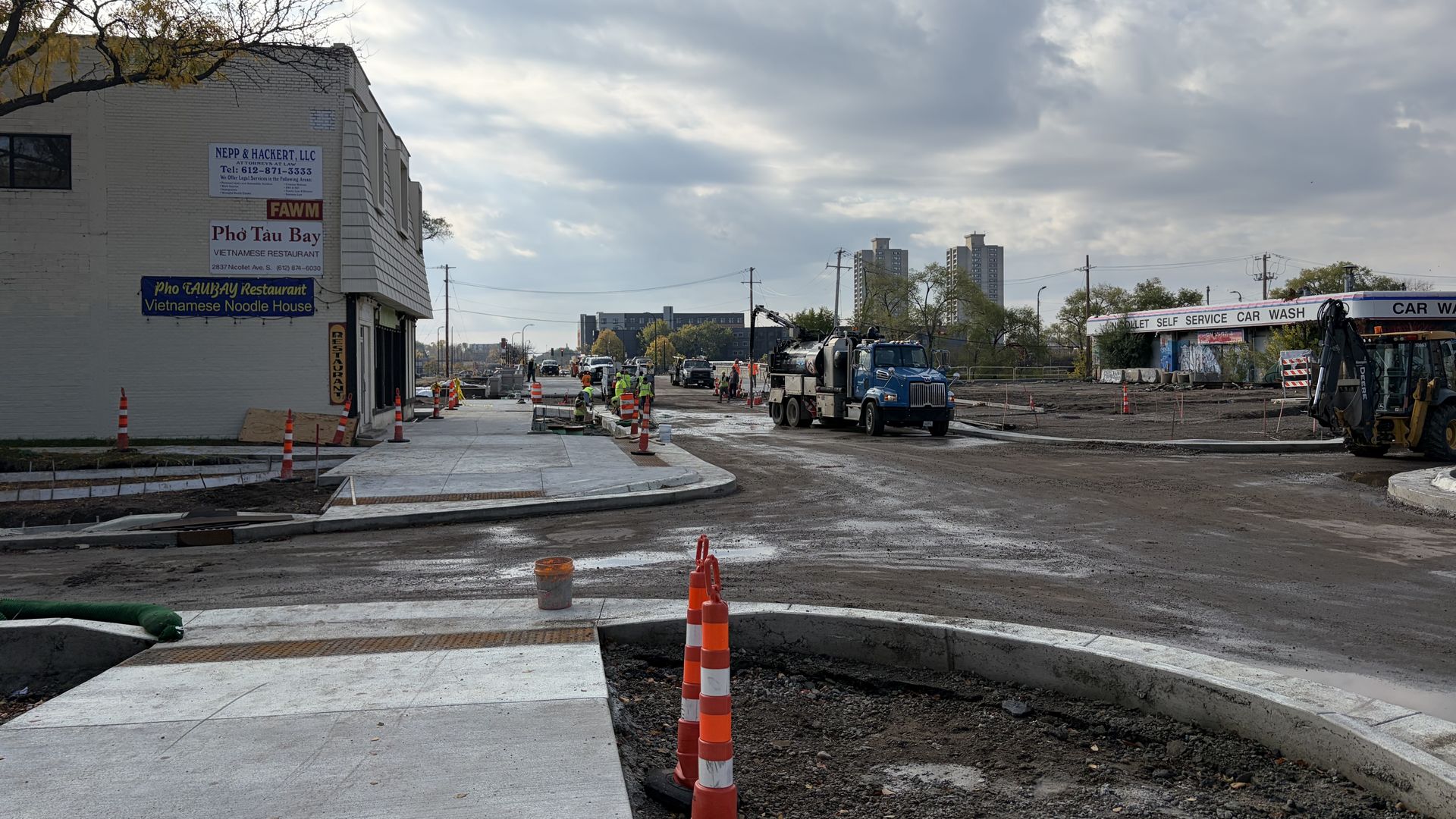 Urban construction scene: left, a white brick building with restaurant signs; orange safety cones line a wet street; construction trucks and machinery on the road; gray cloudy sky overhead.