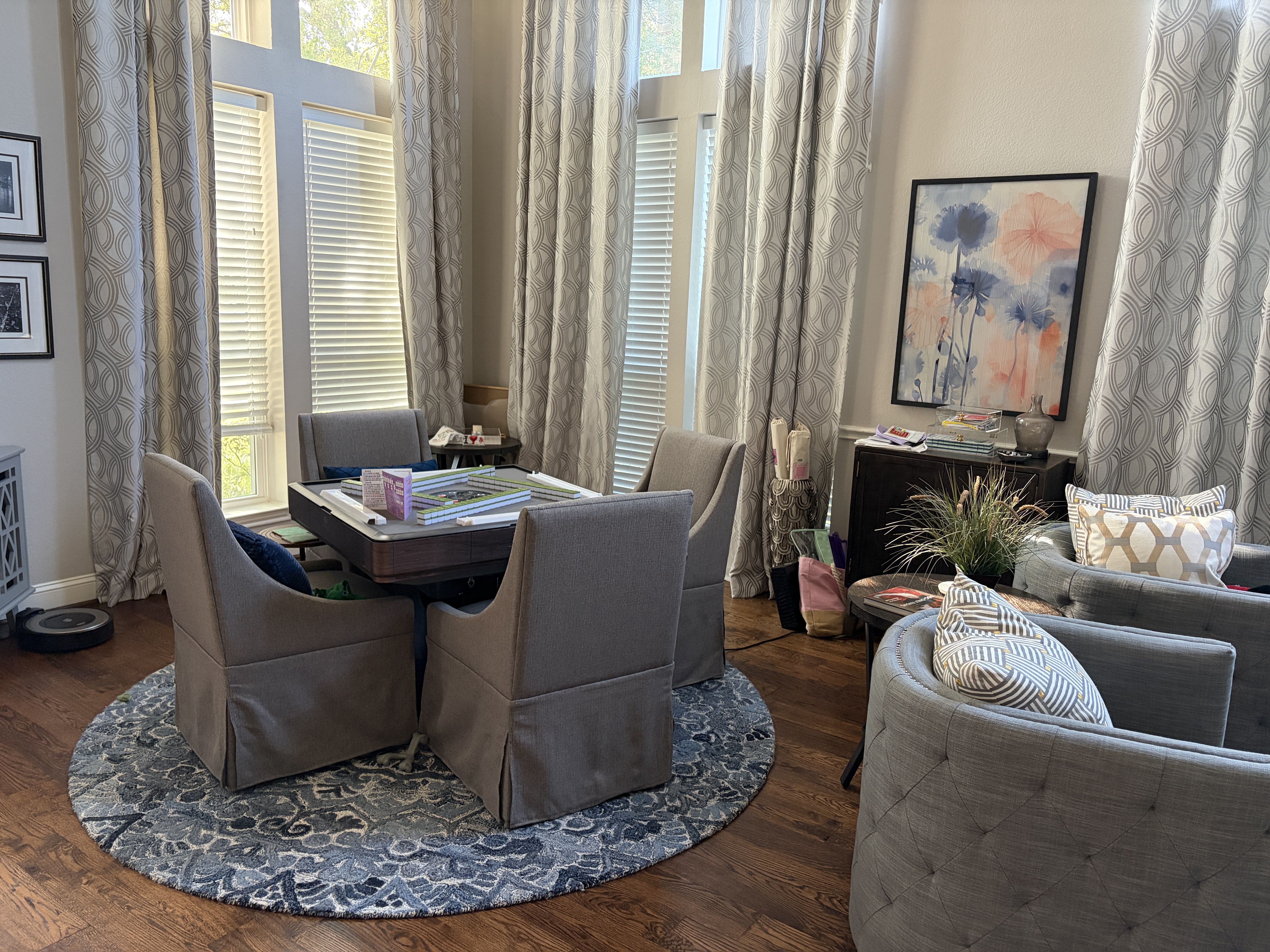 Living room with tall gray patterned curtains on large windowd, a blue circular rug under an automatic mahjong table, gray chairs, and a watercolor floral print above a dark console.