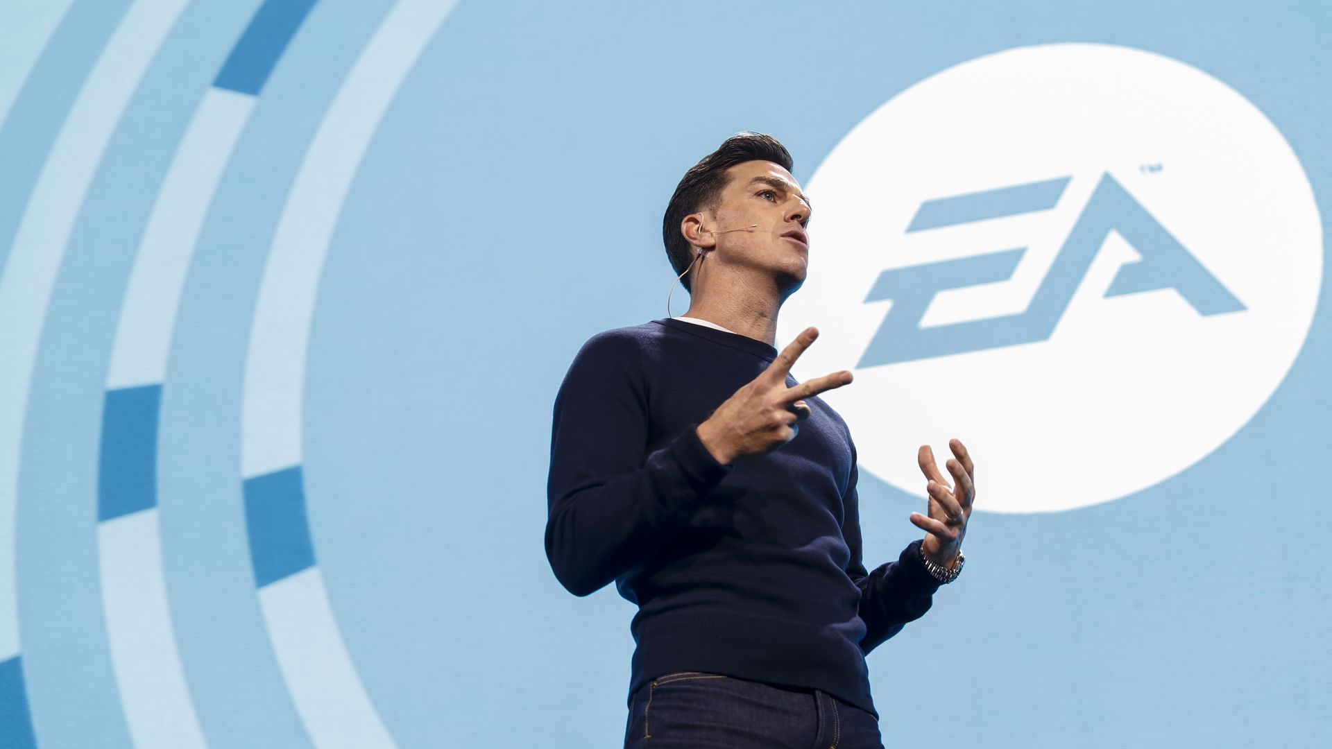 Photo of EA CEO Andrew Wilson in jeans and sweater standing in front of an EA logo