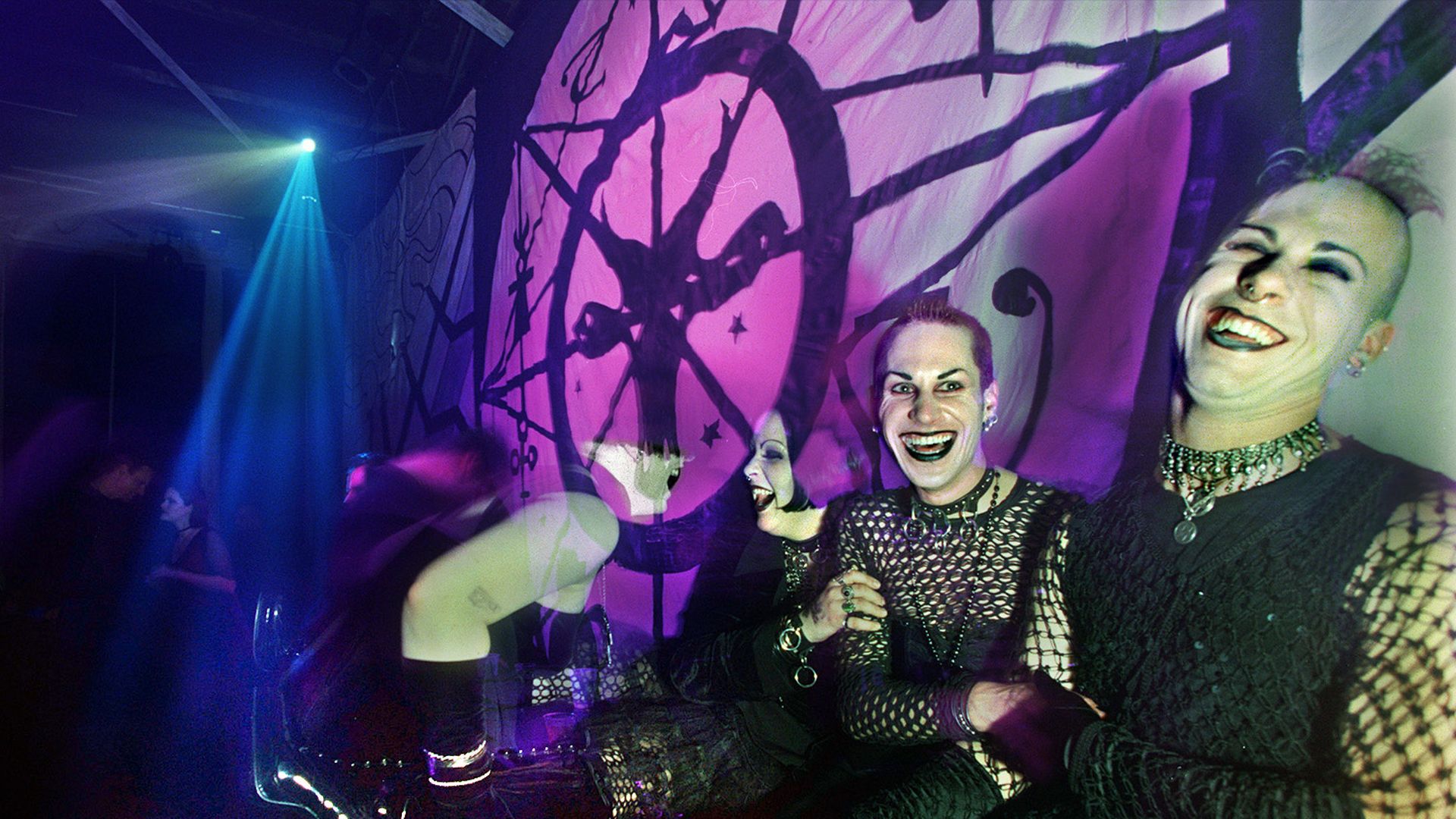 Three people in black mesh outfits laugh and pose at a nightclub; purple-pink lighting bathes the scene, with a graffiti wall showing circular tree motifs and a blue spotlight from the left.