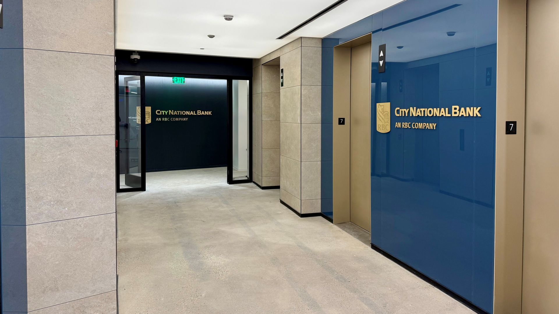 Modern bank lobby corridor with two gold elevators on the right, blue wall panels, and City National Bank logo in gold with "AN RBC COMPANY" underneath on both wall and door at end.
