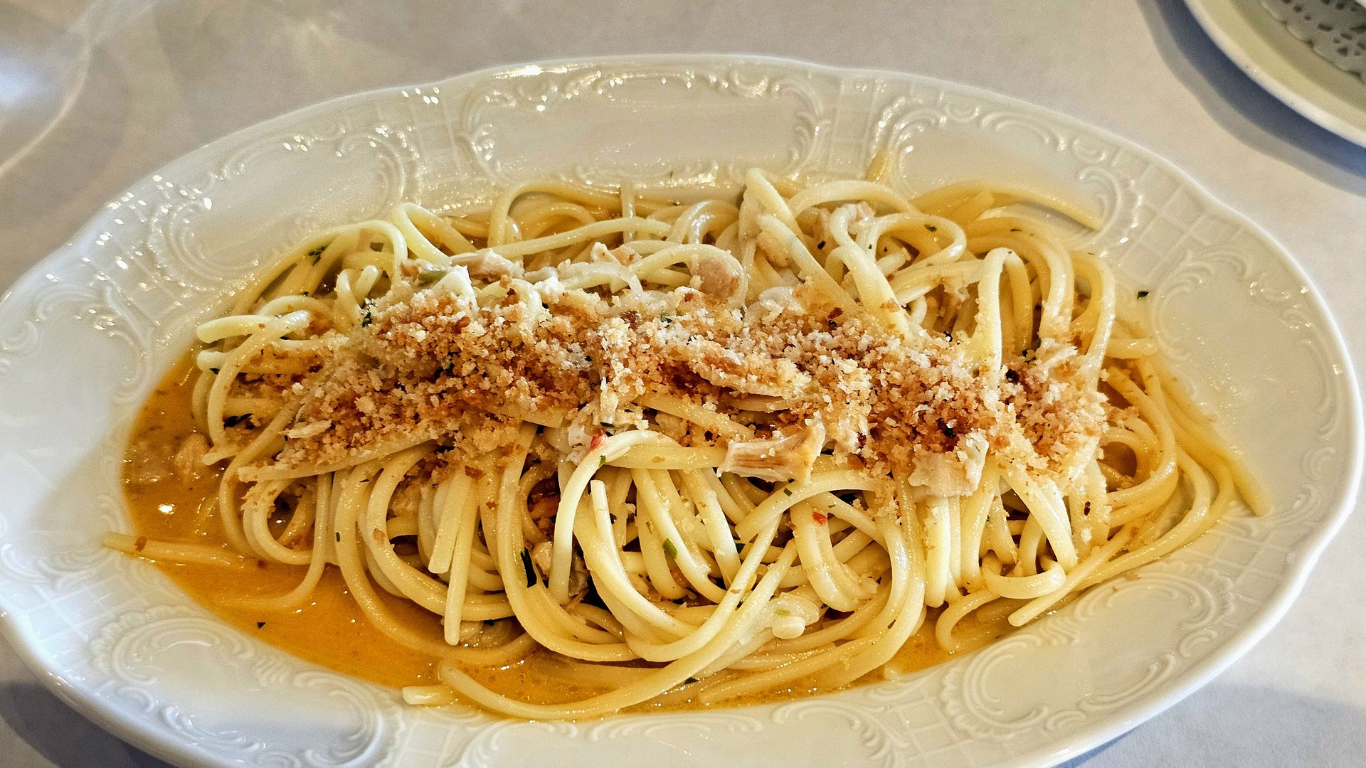 Close-up of a white ornate plate of spaghetti topped with breadcrumbs and grated cheese over a light orange sauce, with specks of parsley.