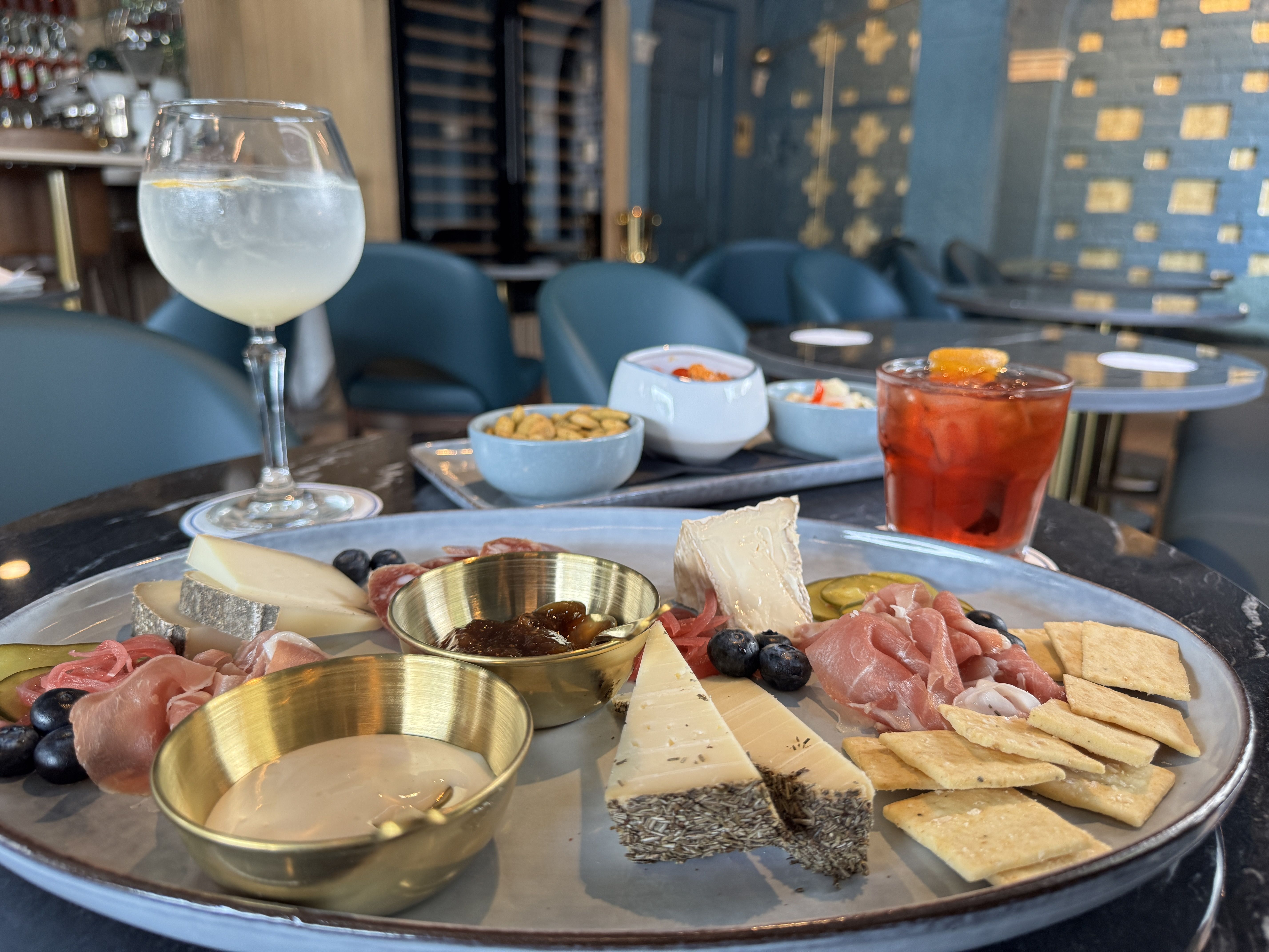 Cheese and charcuterie platter with crackers, pickles, blueberries, and sauces on a metal tray at a modern restaurant table with a cocktail and a glass of water nearby.