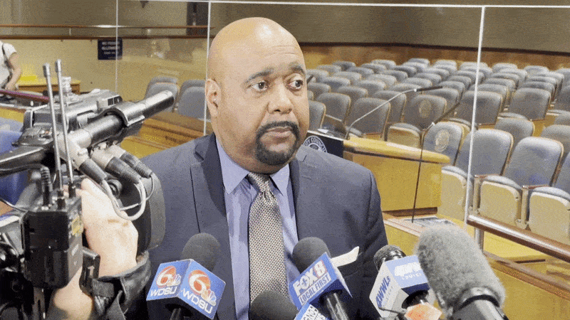 A bald man in a blue suit and lavender shirt speaks at a press conference, surrounded by several microphones; empty auditorium seating in the background.