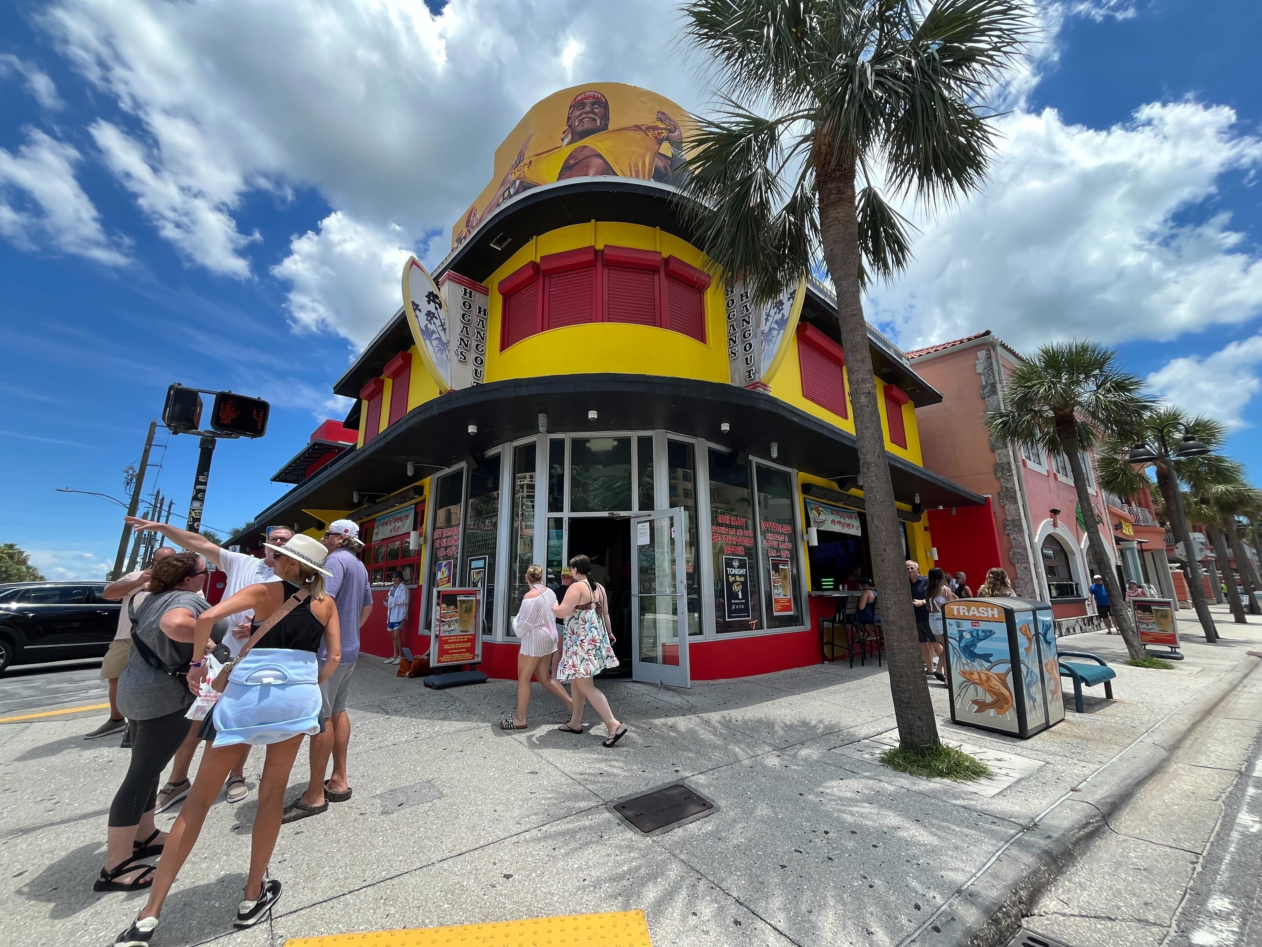 Colorful corner building with yellow and red facade, named Hogan's Hangout, under a blue sky with white clouds, people gathered outside and palm trees lining the sidewalk.
