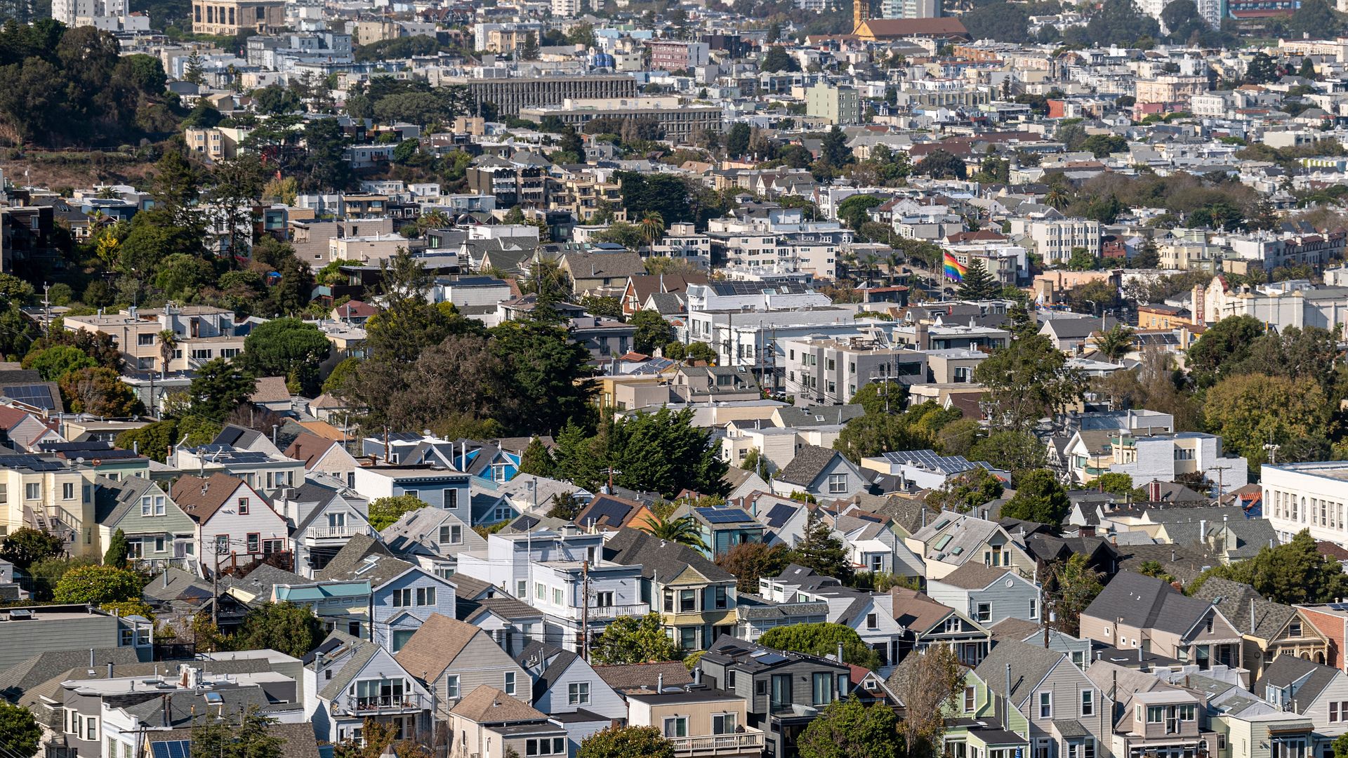 Panoramic view of a dense residential hillside with colorful houses and trees; a rainbow Pride flag is visible on a flagpole among the homes.