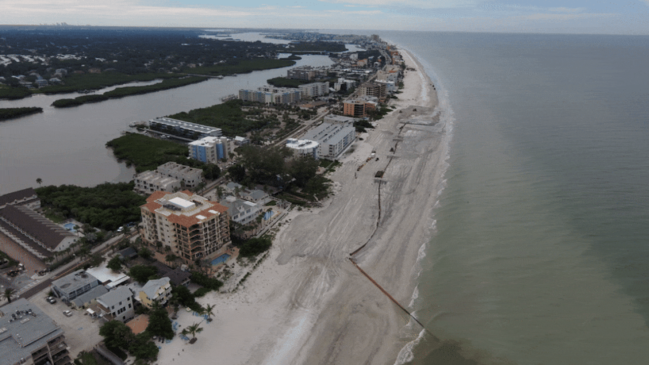 Aerial view of a coastal city with sandy beach, buildings along the shoreline, and construction equipment on the beach. Calm ocean on the right and an inlet with greenery on the left.
