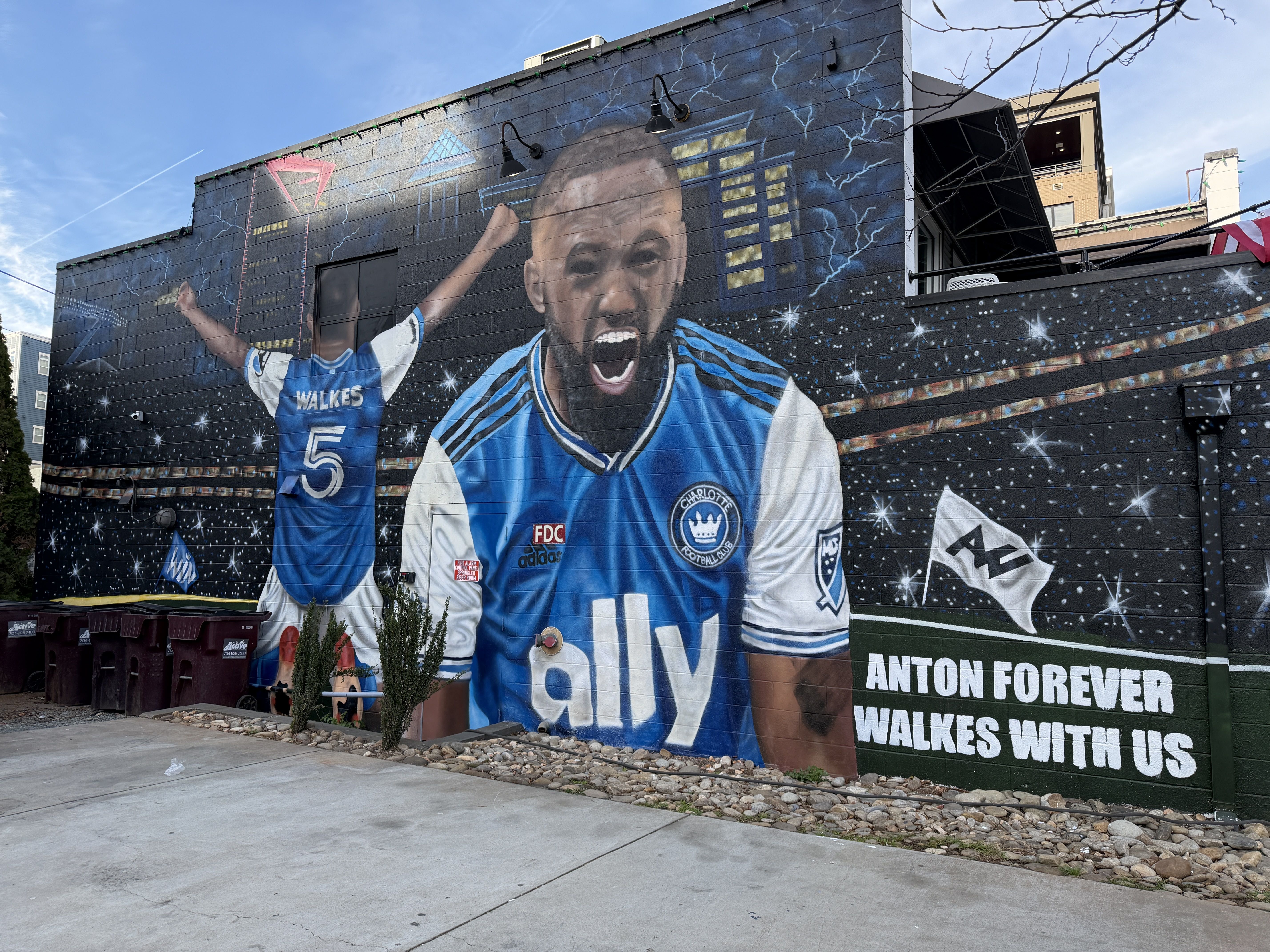 Mural of soccer player Anton Walkes in blue and white Charlotte FC jersey, celebrating with arms raised and shouting, cityscape and starry sky background, text: "Anton forever Walkes with us"