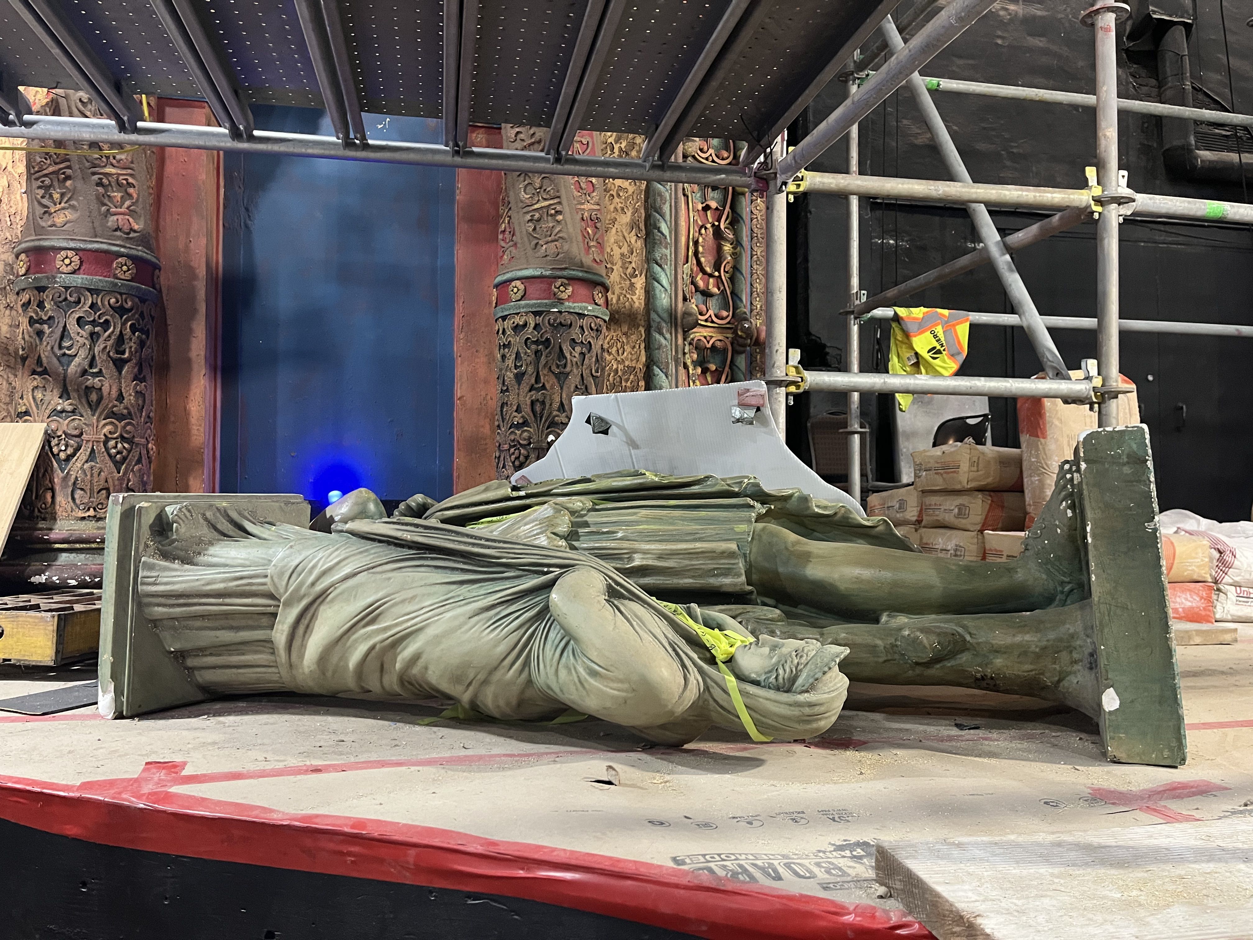 Interior construction scene with ornate carved pillars in red and teal, metal scaffolding, a blue backdrop, scattered tarps and bags, and a yellow safety vest hanging on the frame.